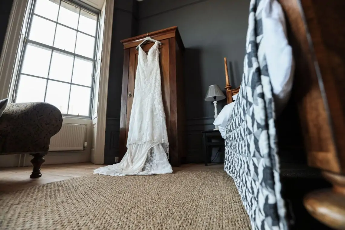 Morilee bridal gown by Madeline Gardner hanging up against wooden wardrobe in a bedroom