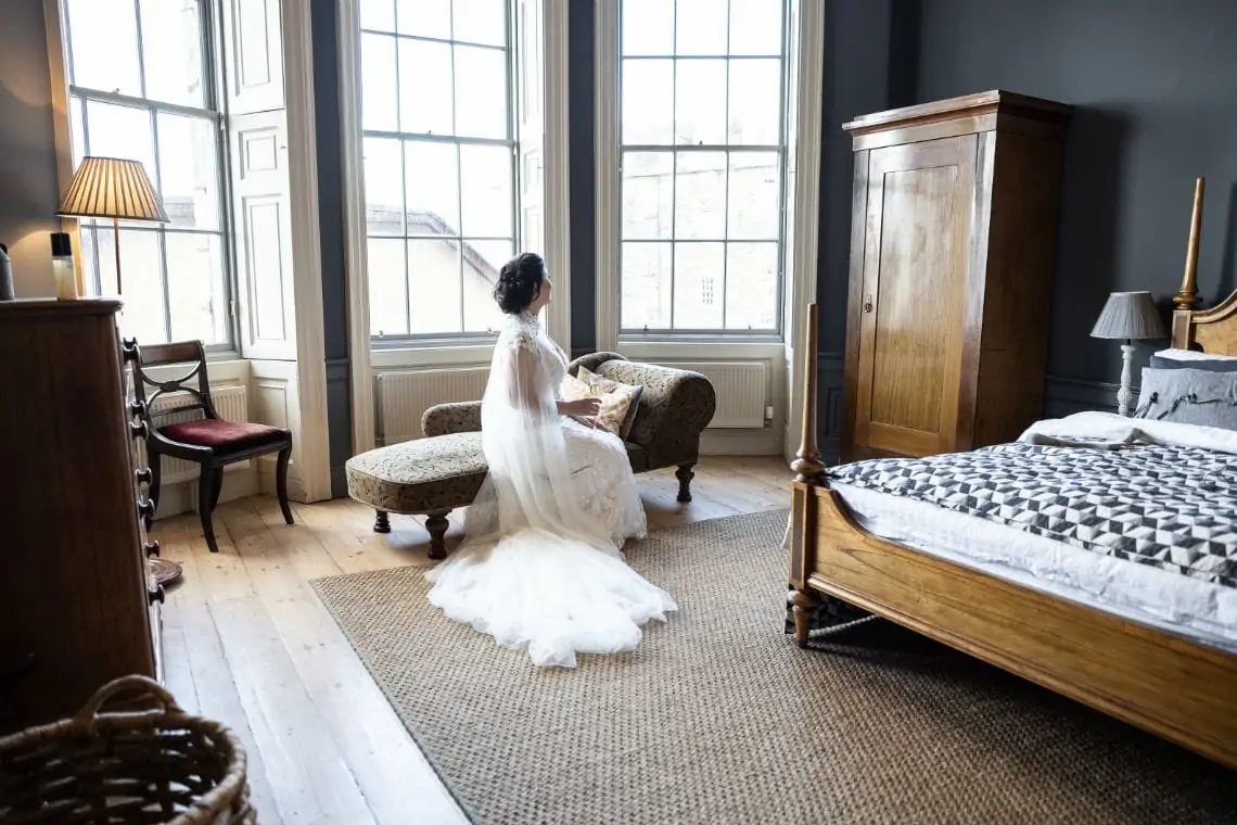 Bride looking out window sitting on chaise longue in bedroom