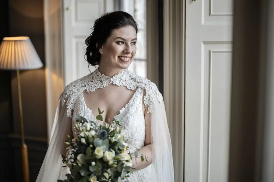 Bride holding bridal flowers looking out of the window and smiling