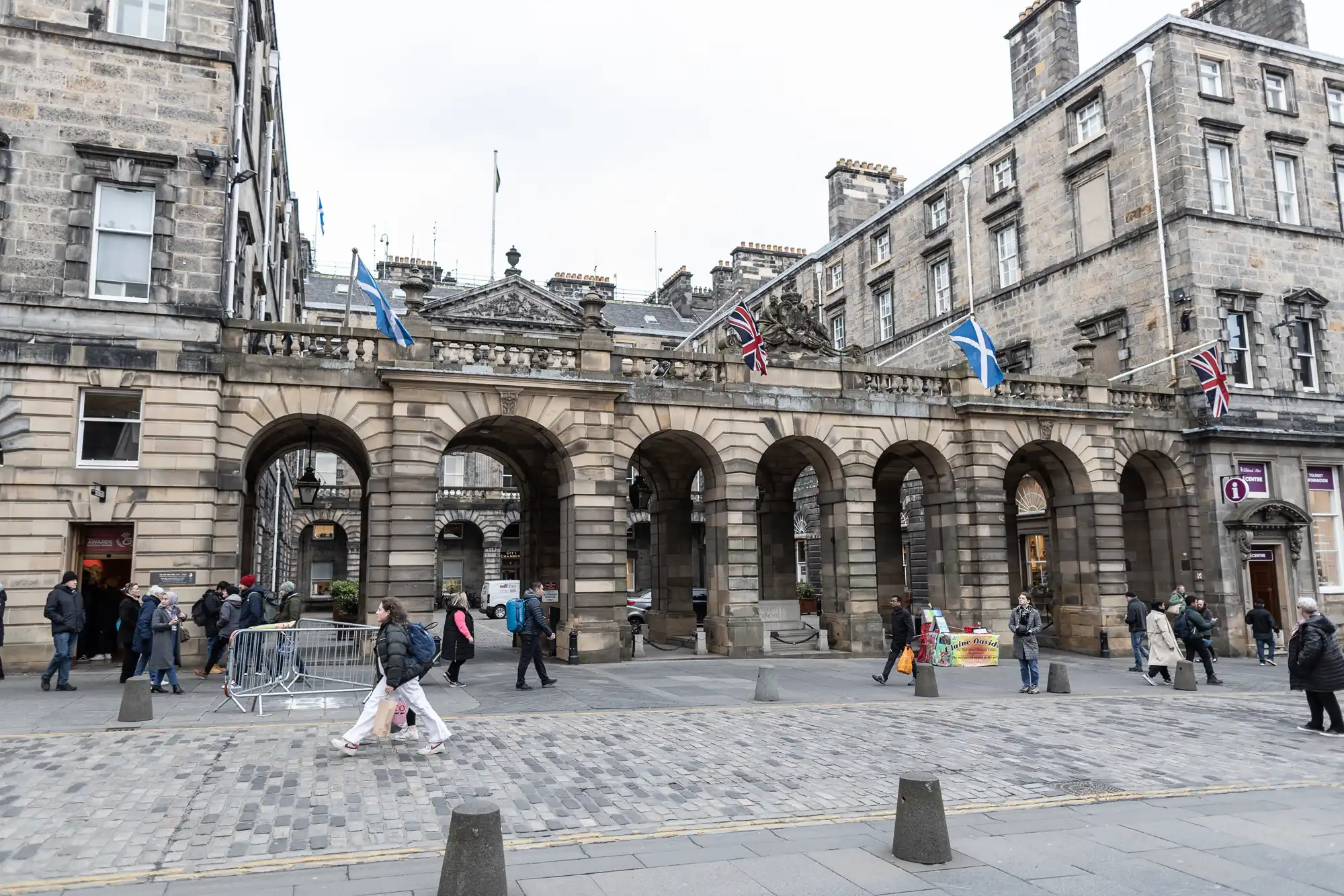 A historic stone building with archways. Several flags hang above, including Scottish and British flags. People walk by and stand in front, with some shopping bags and items visible.