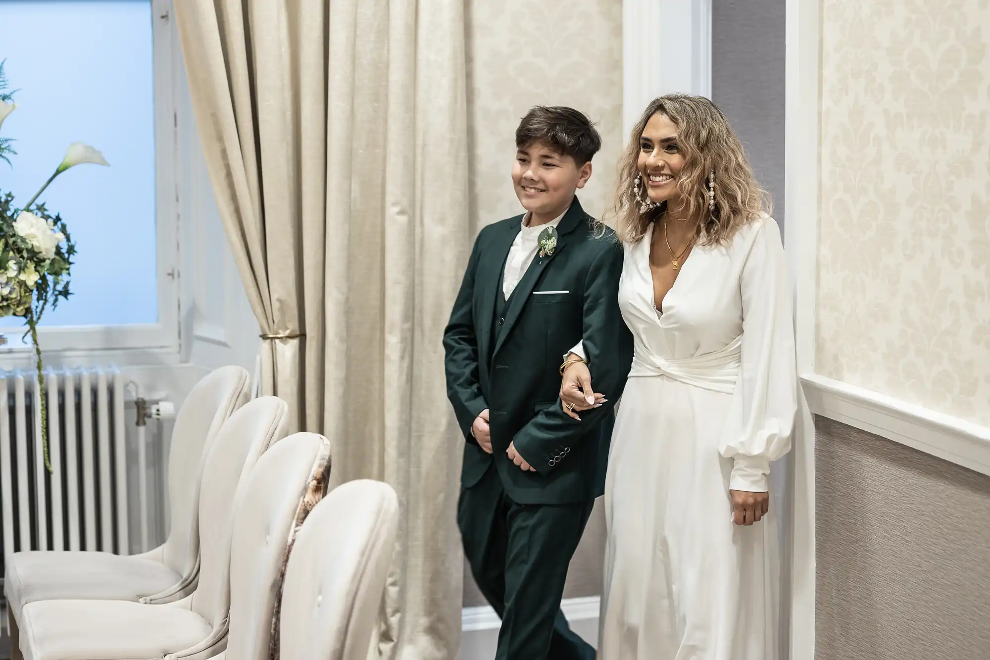 A woman in a white dress and a young person in a dark green suit walk arm in arm through a room with white chairs and beige curtains.