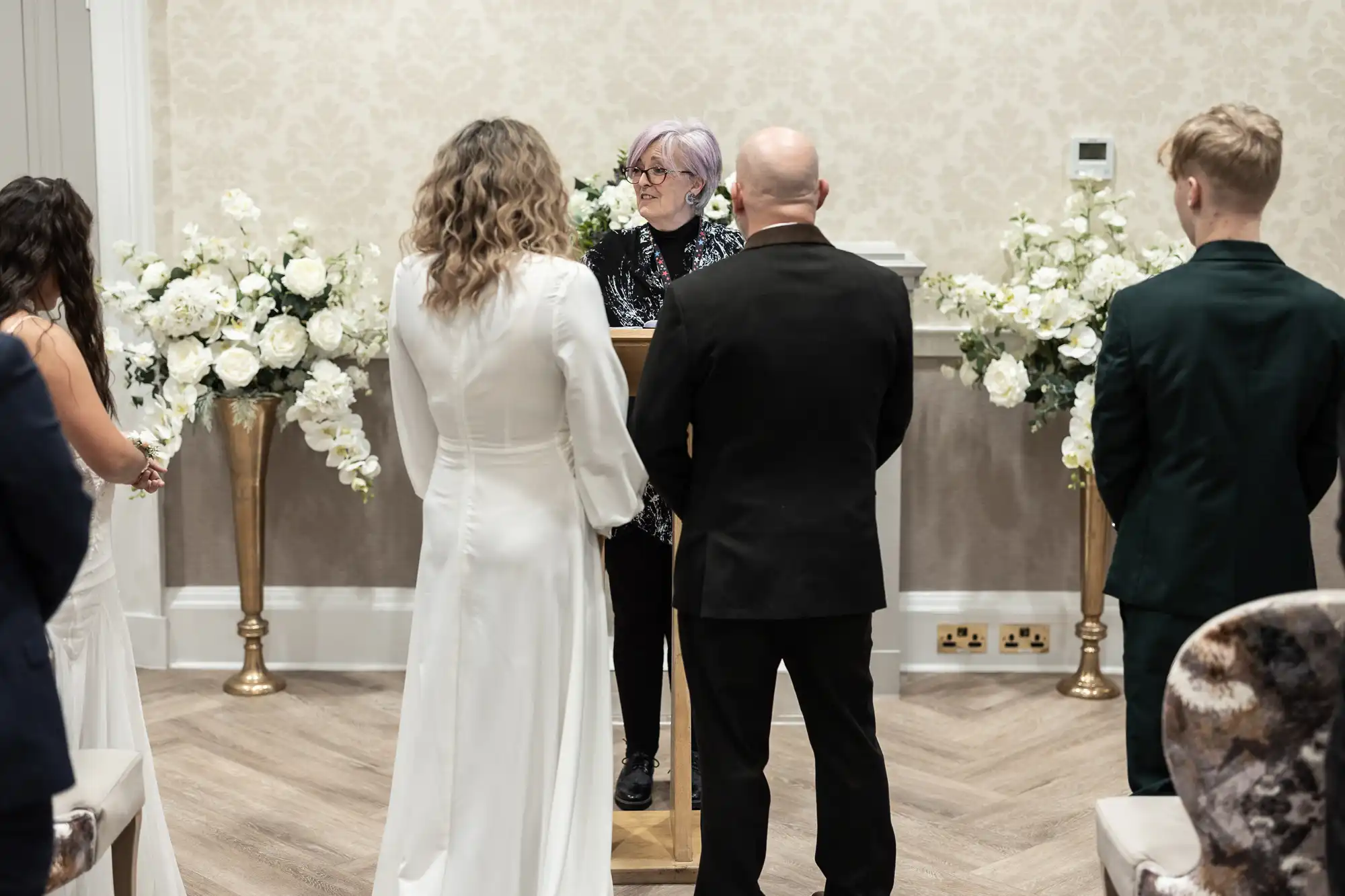 A wedding ceremony with an officiant speaking at a podium. A couple dressed in white and black stand facing the officiant, with two other individuals standing nearby. Flowers adorn the background.