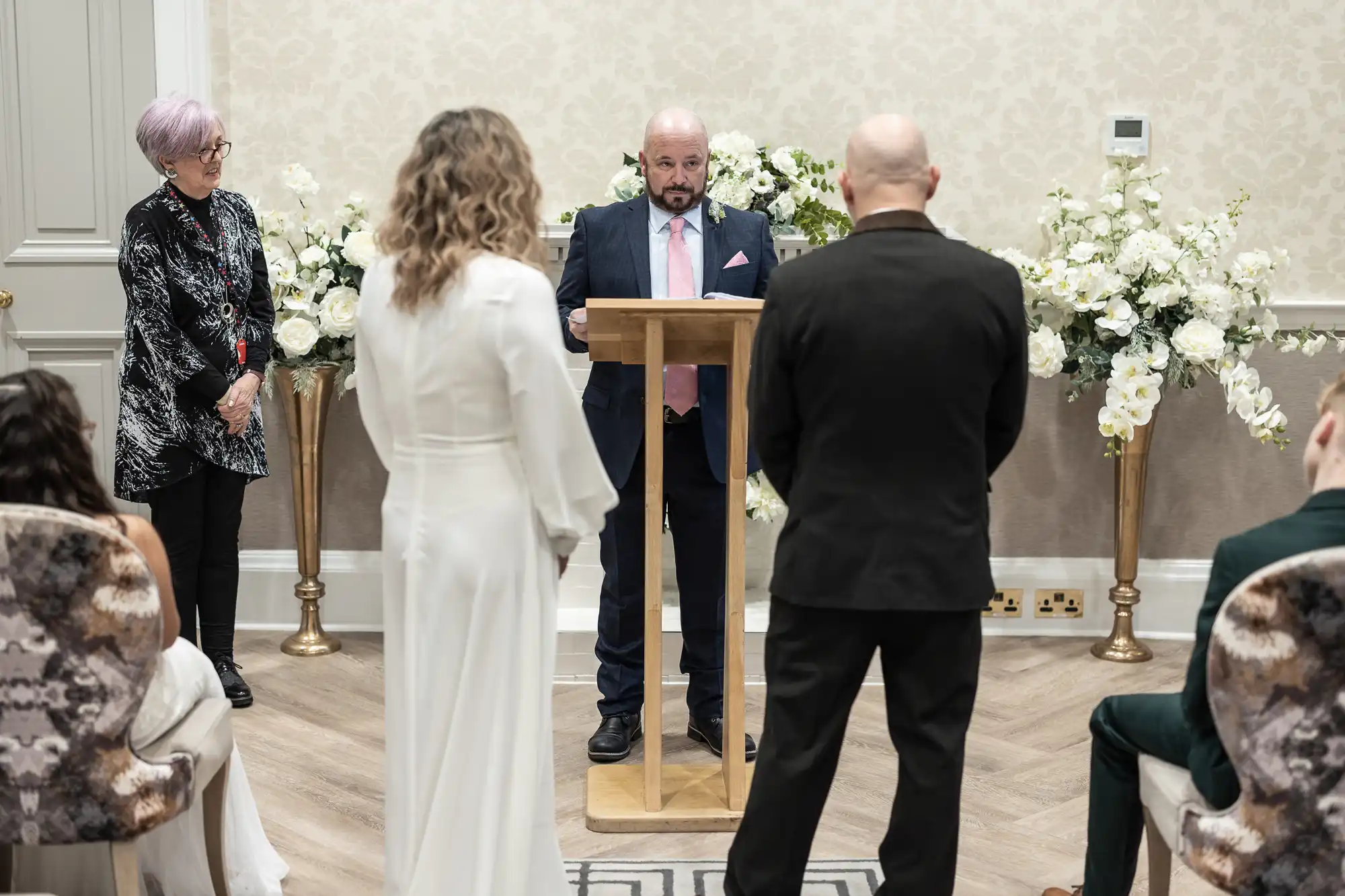 A wedding ceremony in progress with a couple standing facing a person reading from a podium. Two floral arrangements are visible in the background.