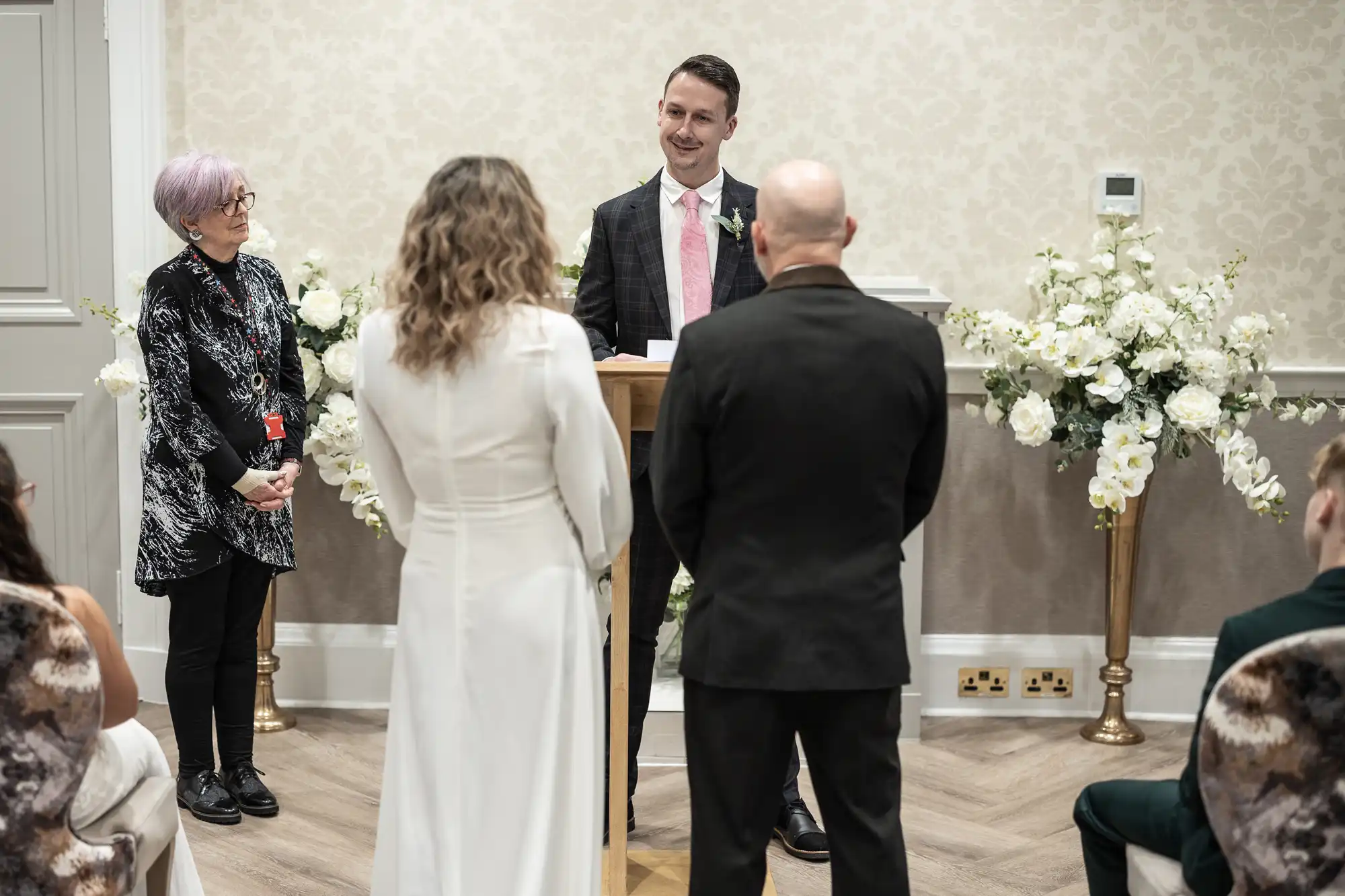 A man stands at a podium speaking to a couple facing him. Another woman stands on the side. Floral arrangements decorate the room, suggesting a formal or ceremonial event.