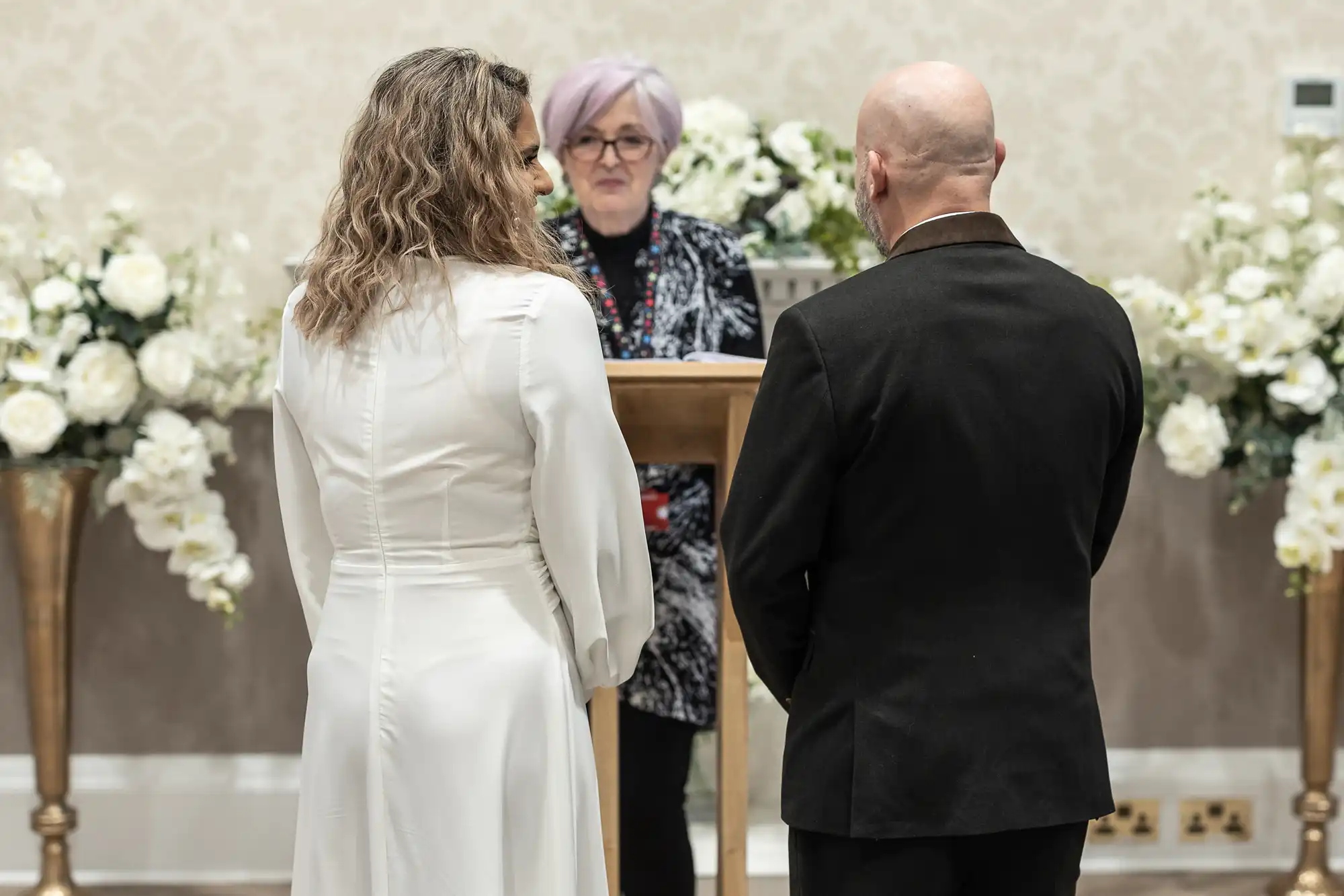 A couple stands in front of an officiant during a wedding ceremony. The officiant is positioned behind a podium, and floral arrangements are seen in the background.
