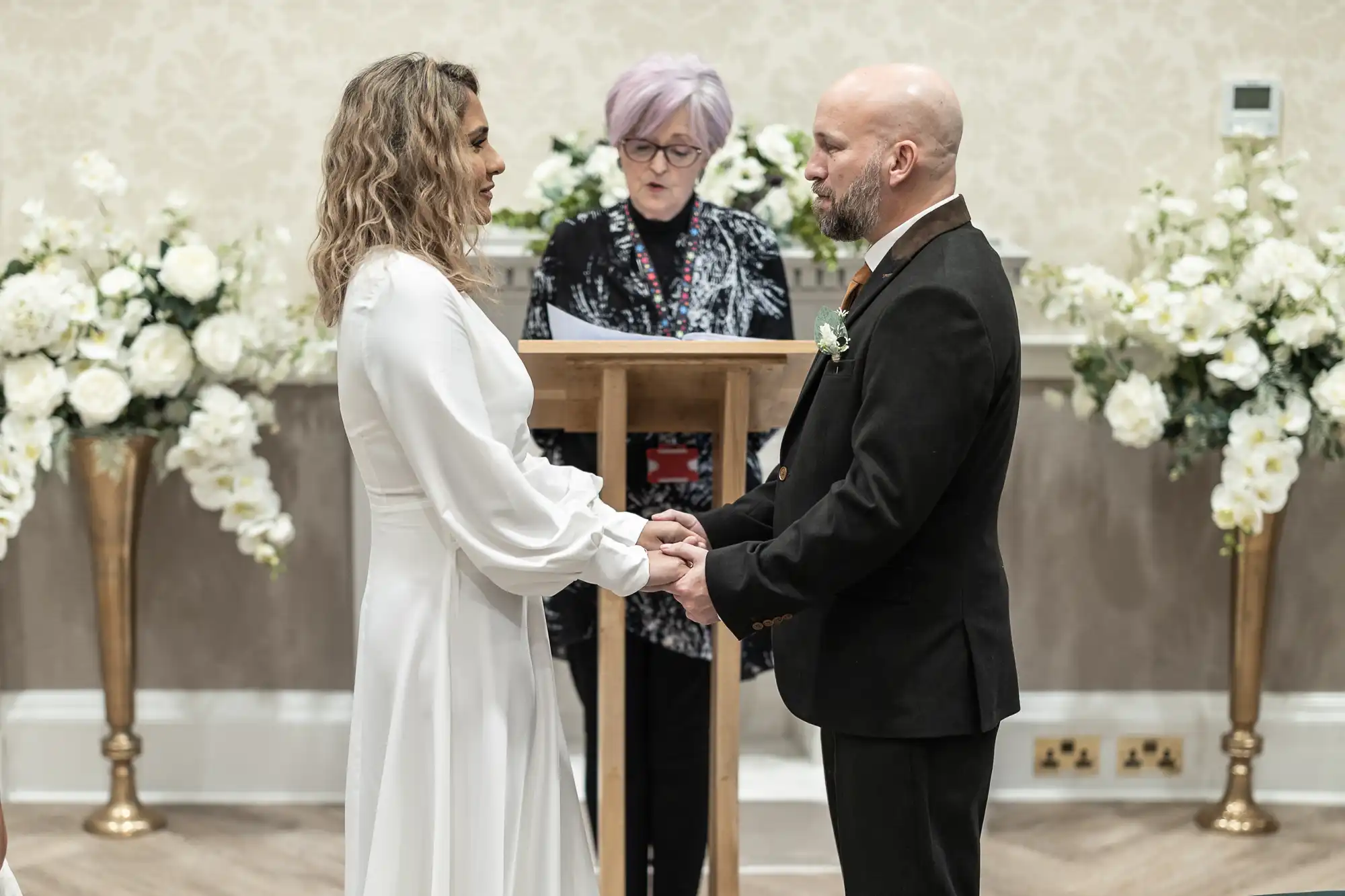 A couple stands facing each other, holding hands, during a wedding ceremony. A person officiates behind them, with arrangements of white flowers on either side.
