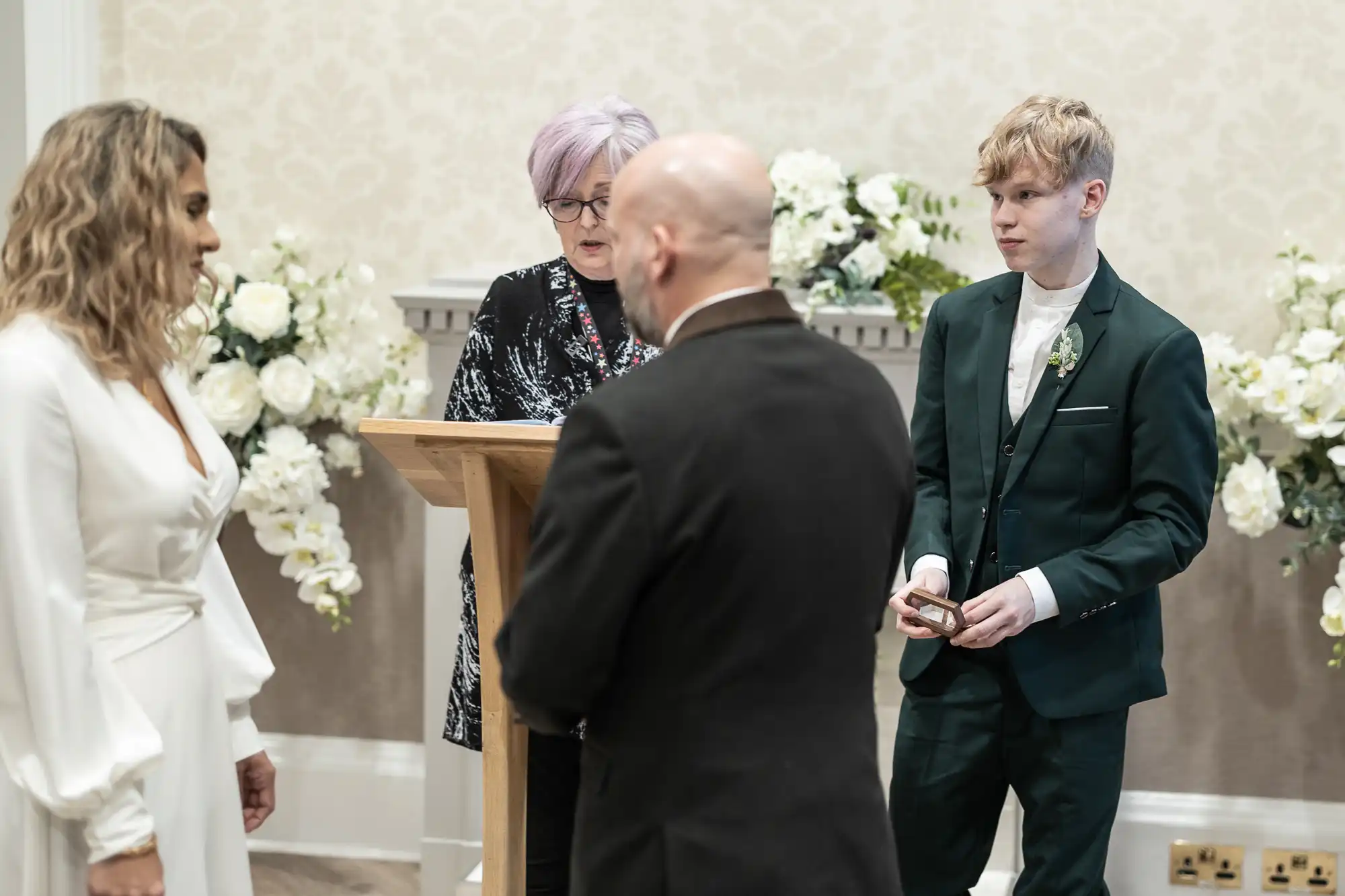 A bride and groom are standing opposite each other in front of an officiant at a wedding ceremony. Another person in a green suit stands nearby holding a small object, likely a ring box.