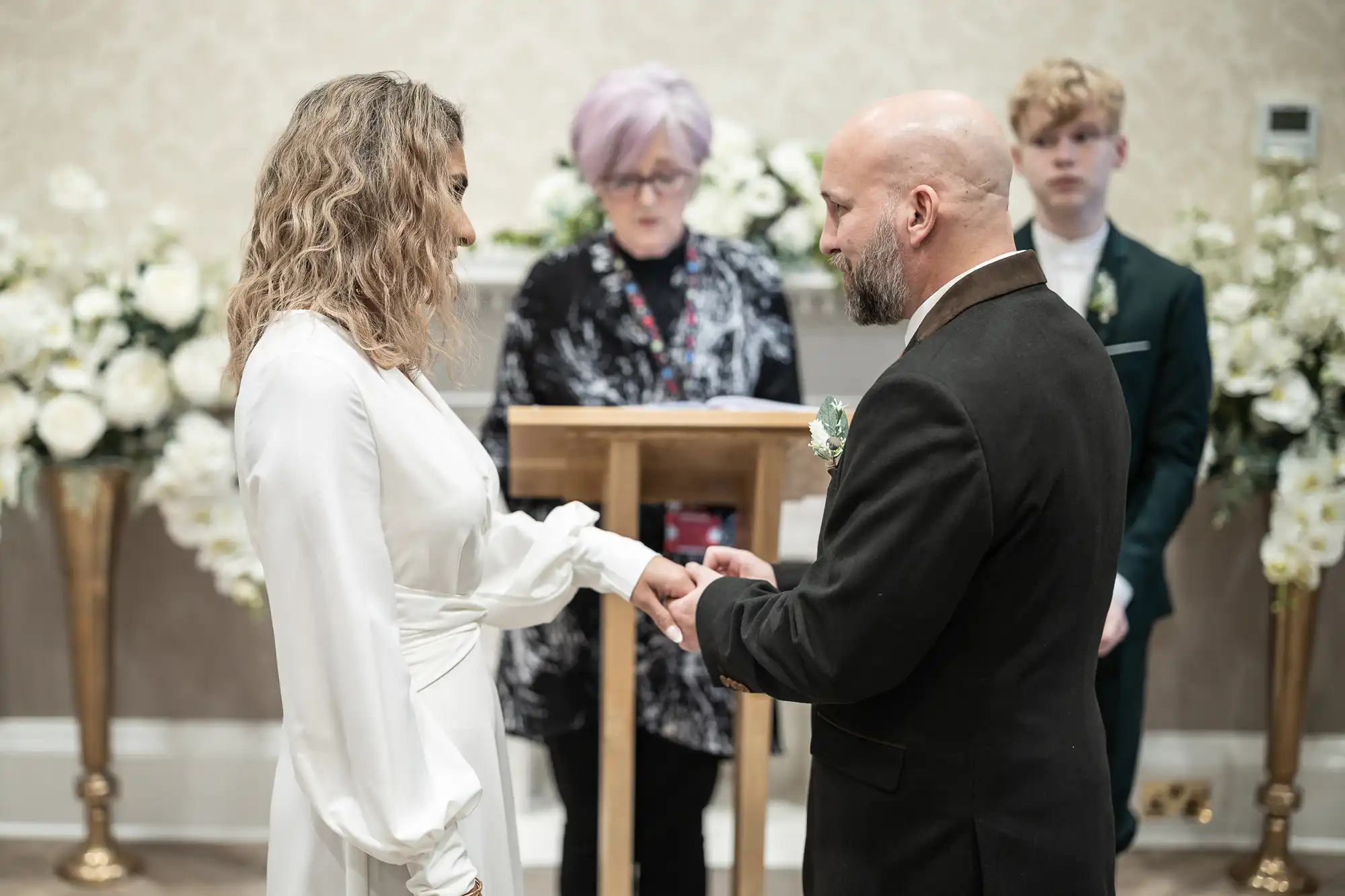 A couple exchanges rings during a wedding ceremony officiated by a person standing at a podium. White floral decorations are in the background. A young person is standing in the background.