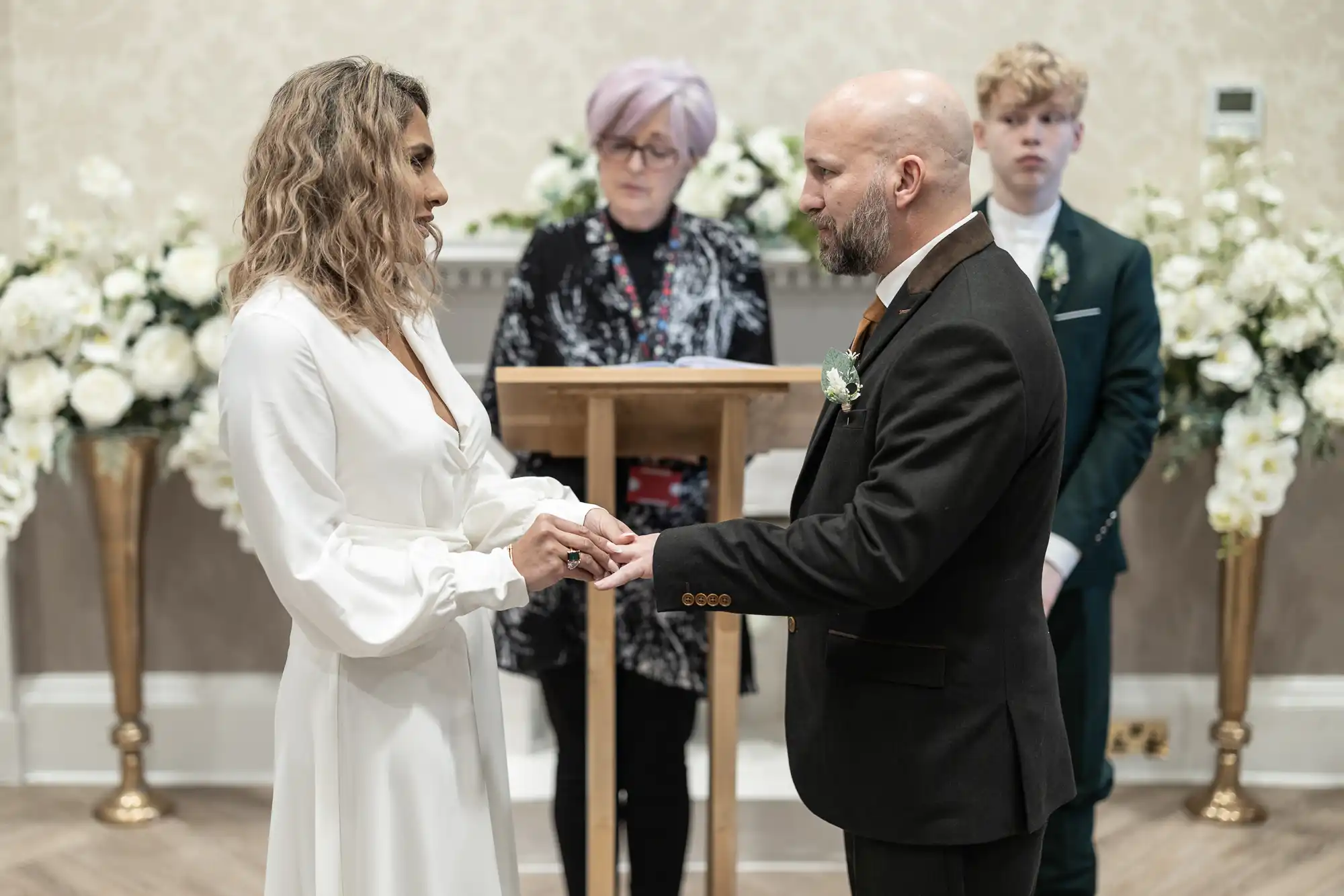 A couple stands exchanging rings during a wedding ceremony, with an officiant and a young boy standing behind them amidst flower arrangements.