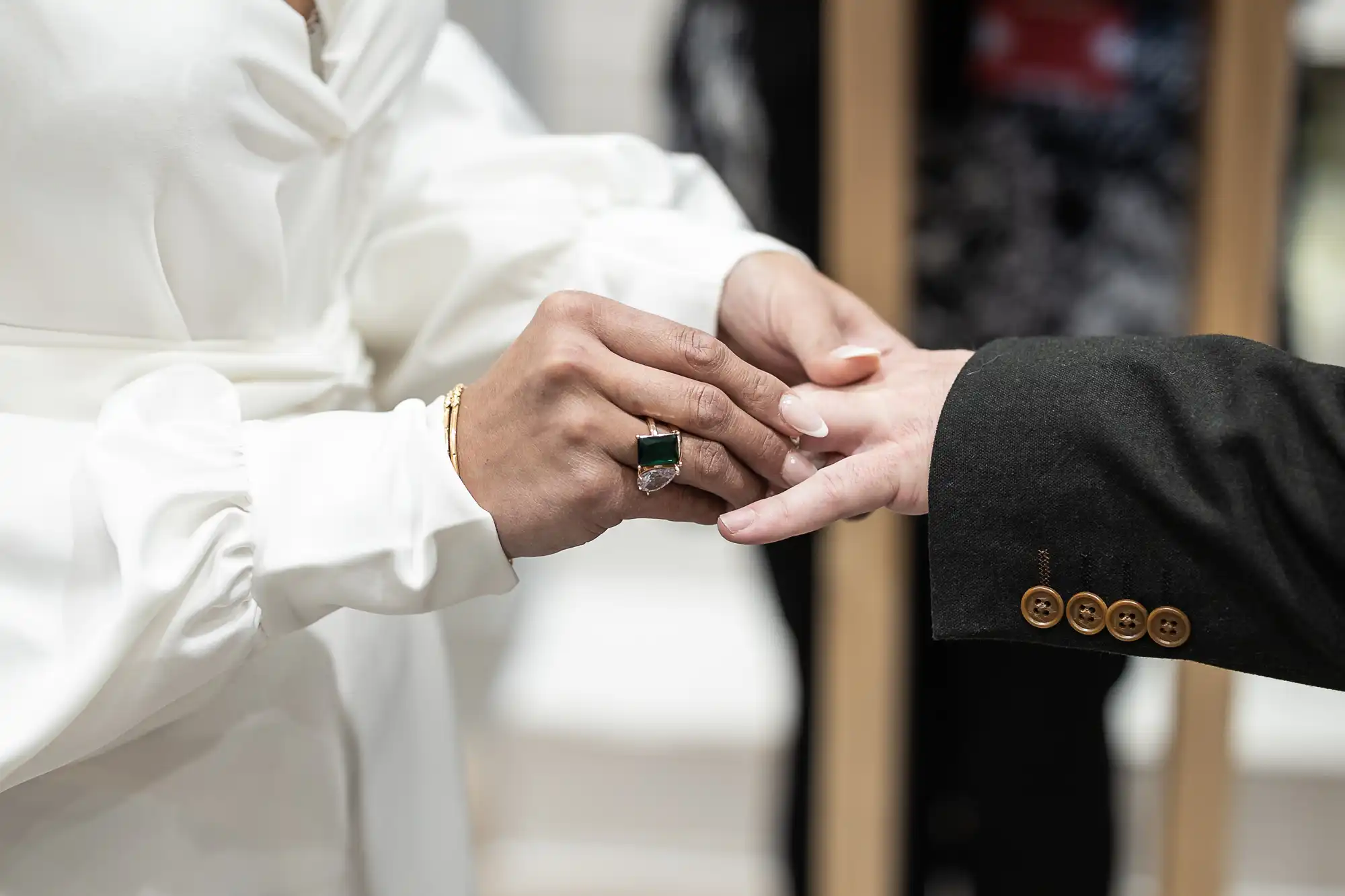 A person in a white garment places a ring on another person's finger, who is wearing a black suit jacket.