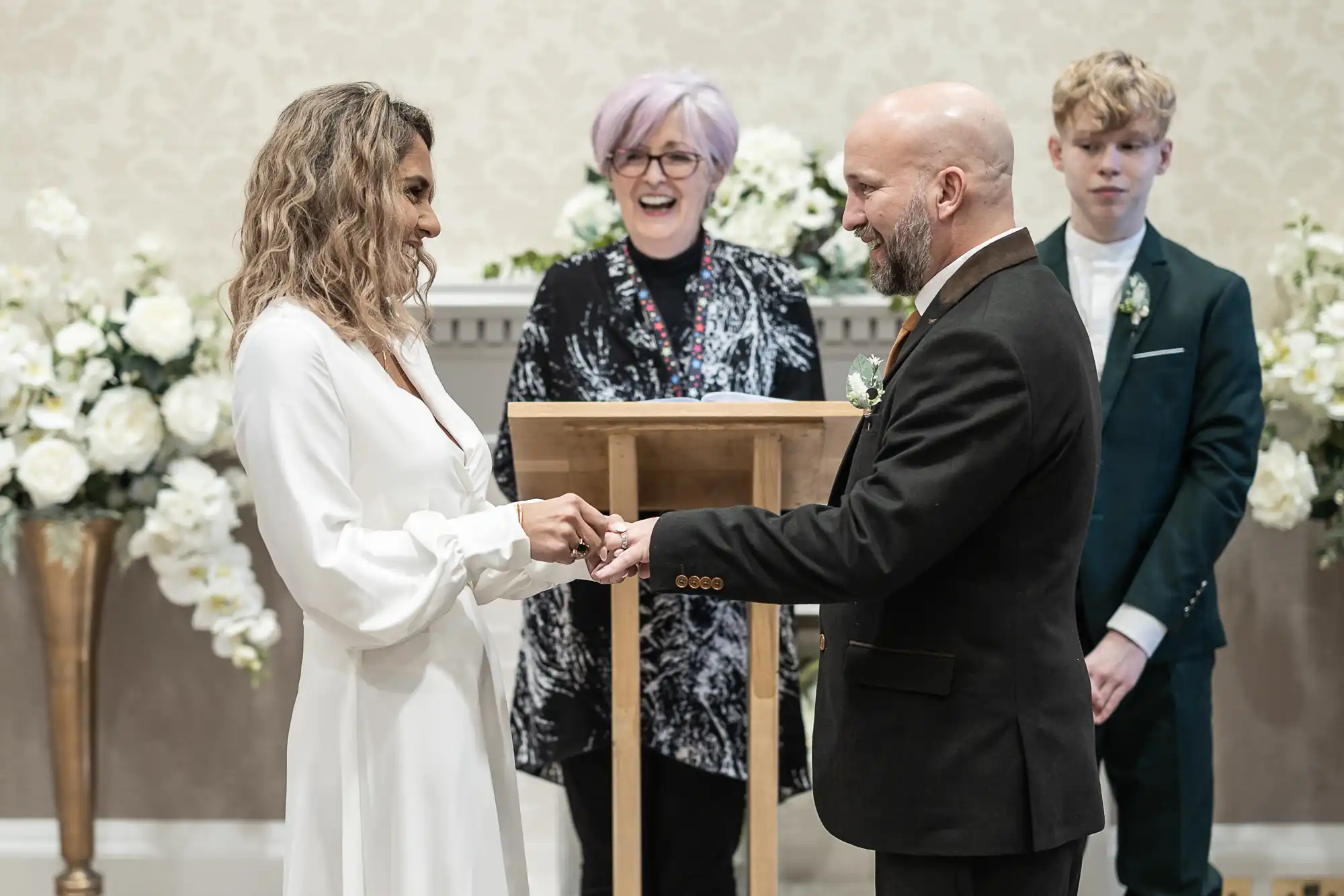 A couple exchanges rings during a wedding ceremony. A woman in a floral dress officiates, and a teenage boy in a suit stands behind them. White floral arrangements decorate the background.