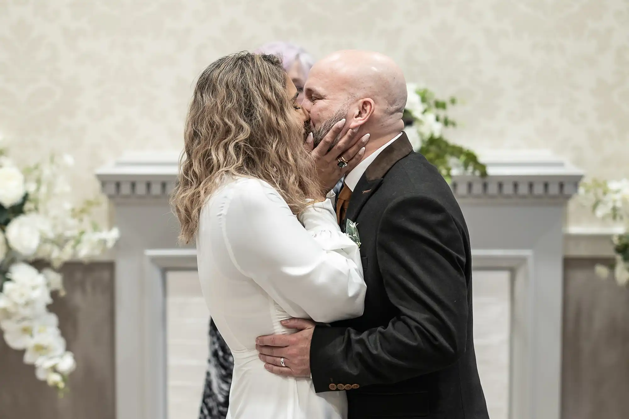 A couple dressed in formal attire kisses during their wedding ceremony, standing in front of a decorated altar with white flowers.