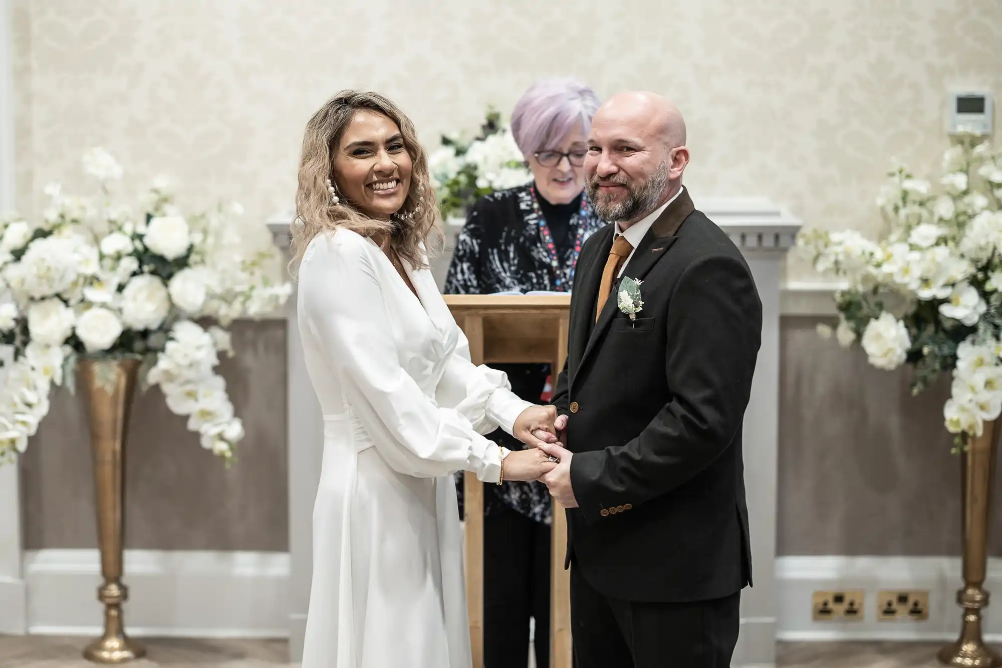 A couple holds hands and smiles at their wedding ceremony, with the officiant standing behind them. There are flower arrangements on either side.