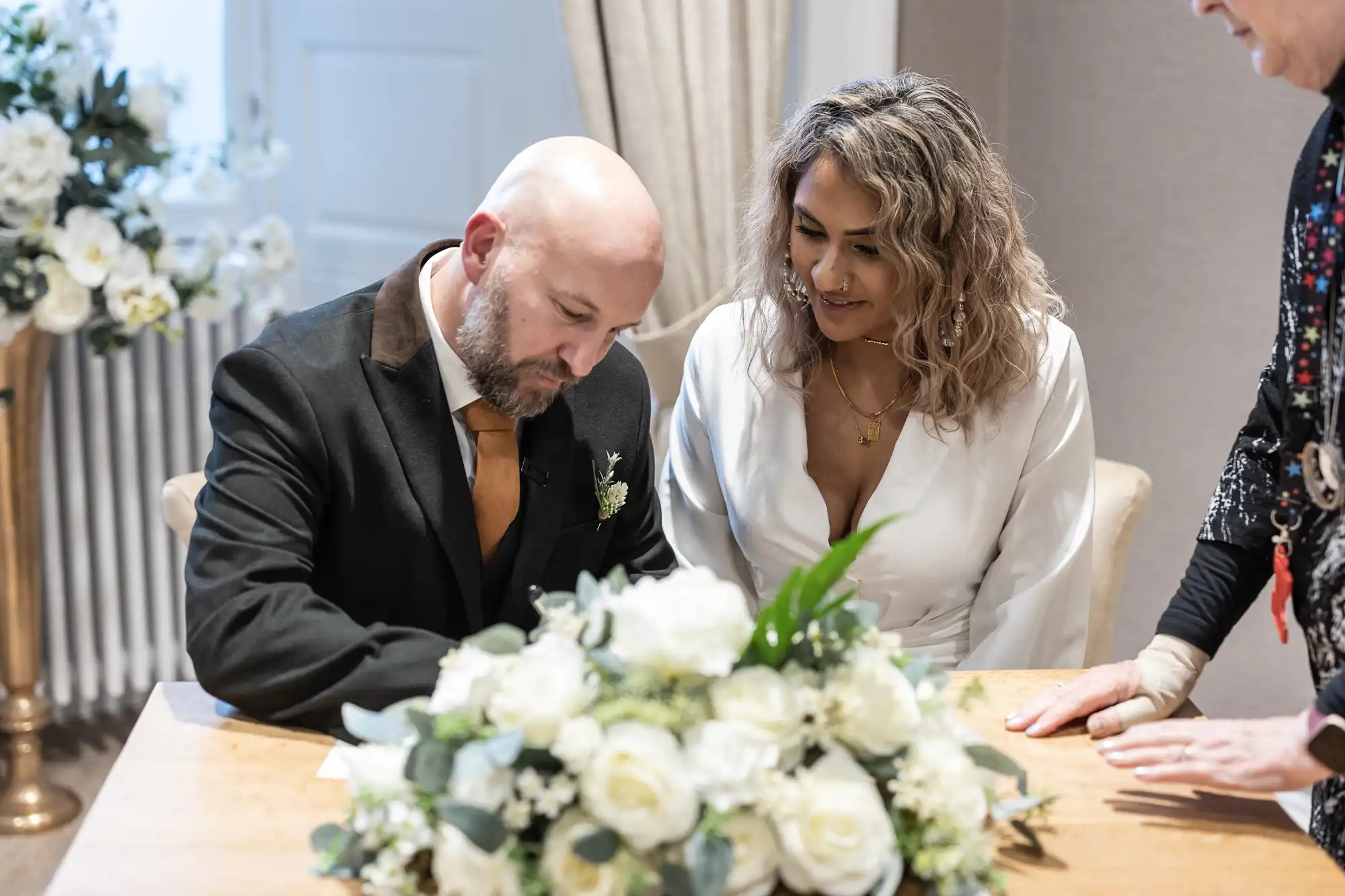 A man and woman are sitting at a table. The man is signing a document while the woman looks on. They are dressed formally, and there is a bouquet of white flowers on the table.