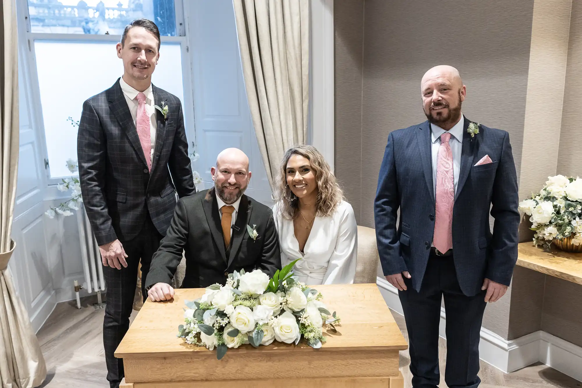 Four people dressed in formal attire pose around a small wooden table with a floral arrangement in what appears to be a wedding ceremony setting.