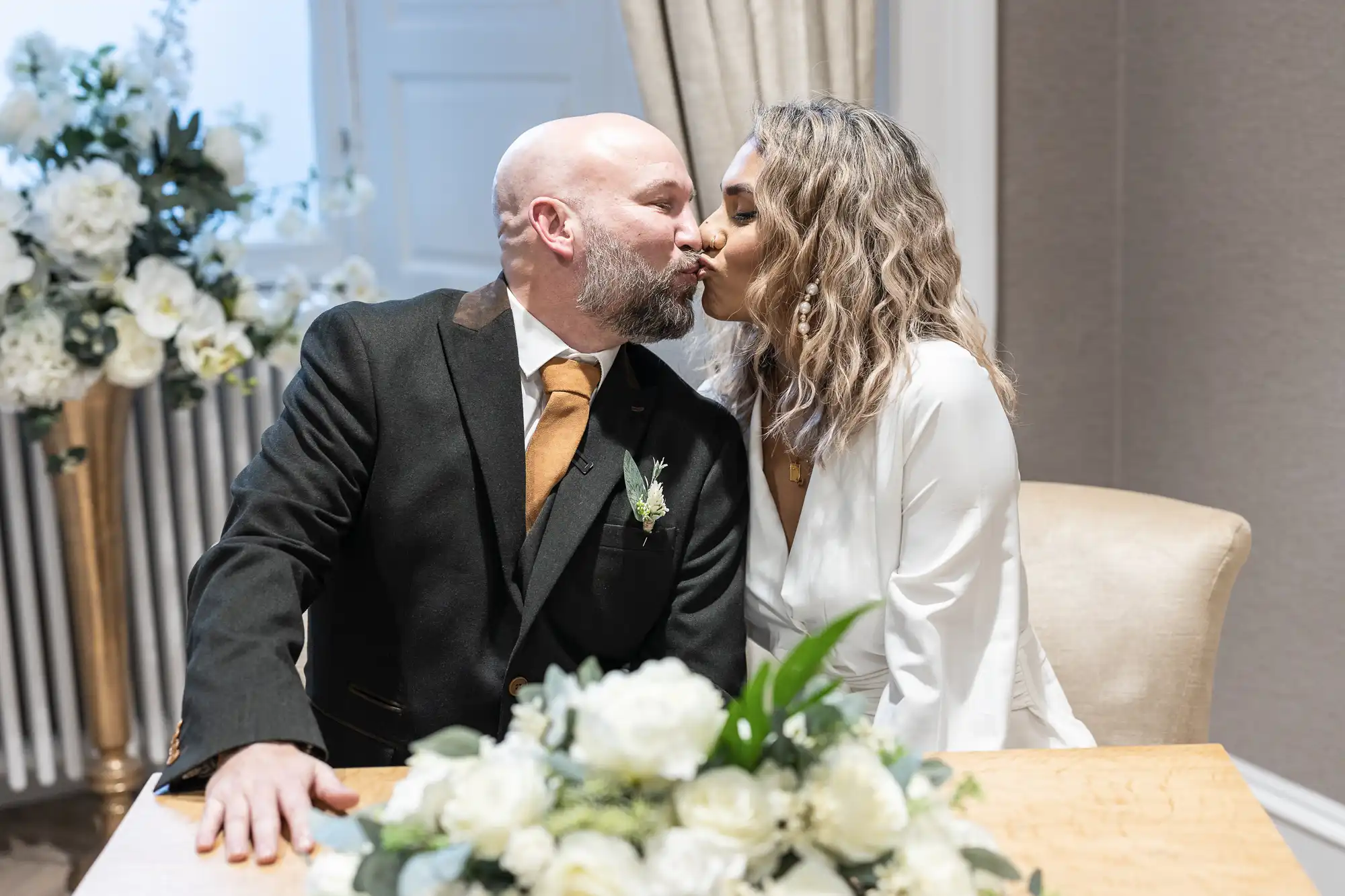 A bride and groom kiss while seated at a table with white floral arrangements.