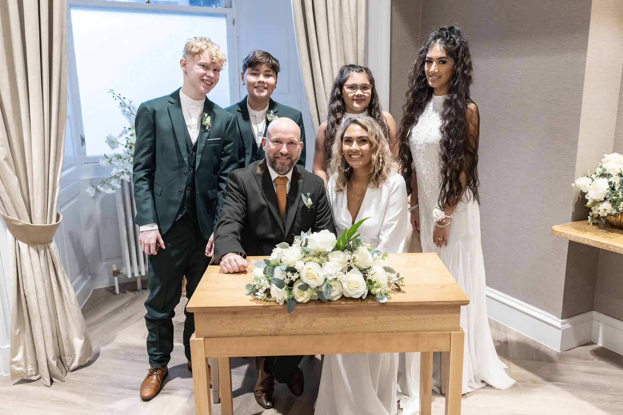 A group of six people posing for a photo in a room with a desk adorned with white flowers. A seated couple, presumably newlyweds, are surrounded by four individuals standing behind them.