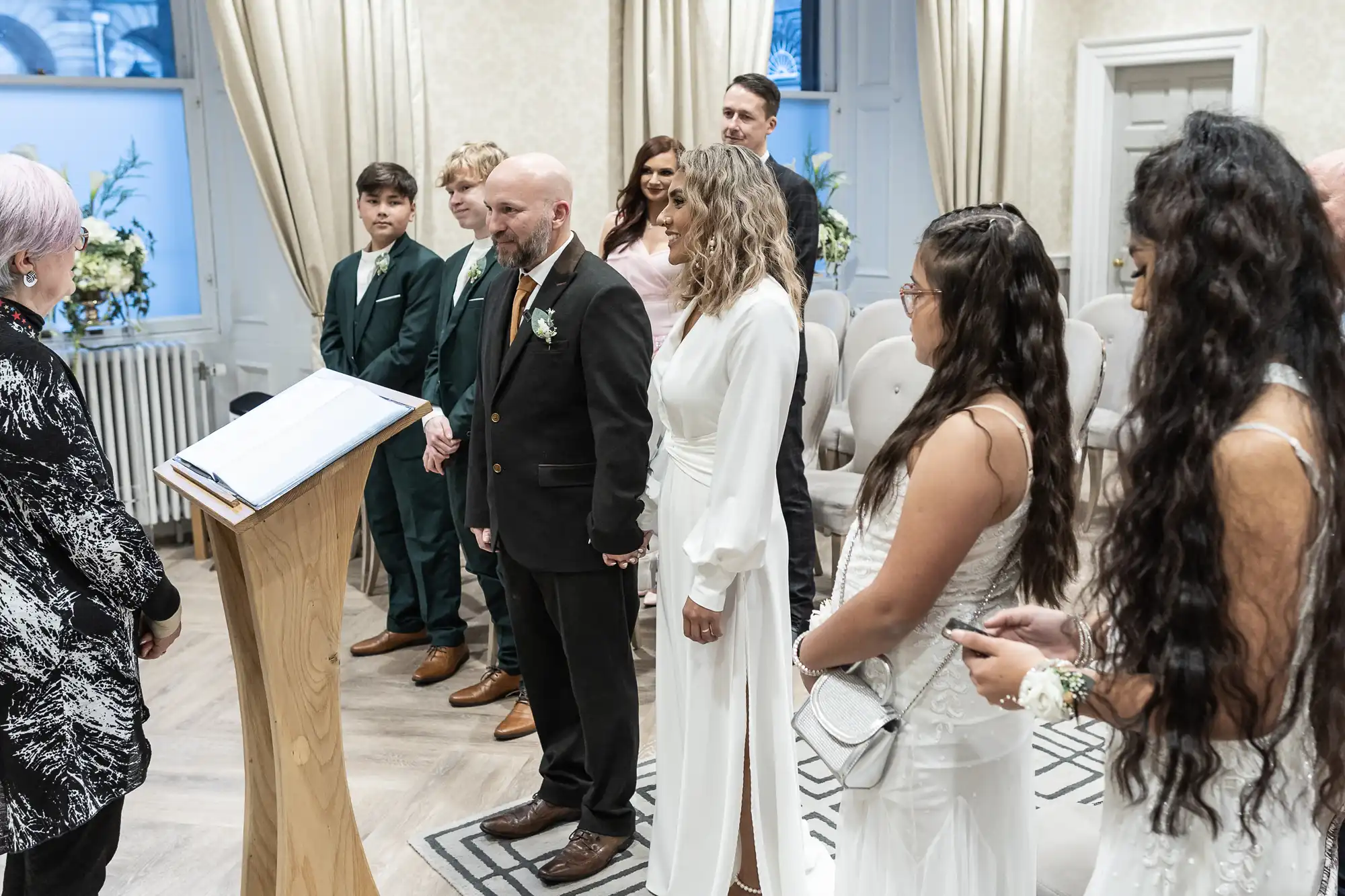 A wedding ceremony with a bride in a white dress and a groom in a black suit, standing with others, facing an officiant at a podium in a small indoor venue.