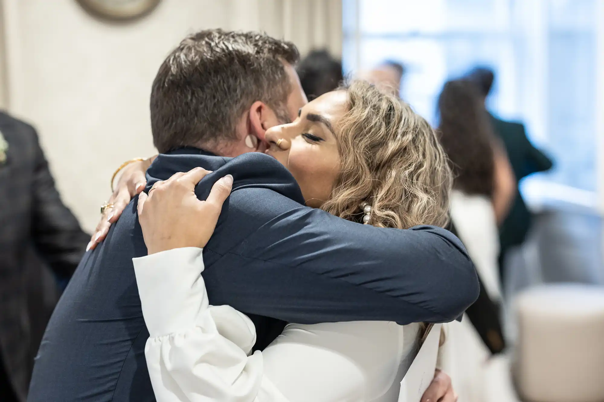 Two people are embracing in a hug at what appears to be an indoor social event. The woman has curly hair and is wearing a white outfit, while the man is dressed in a dark suit. Other attendees are visible in the background.