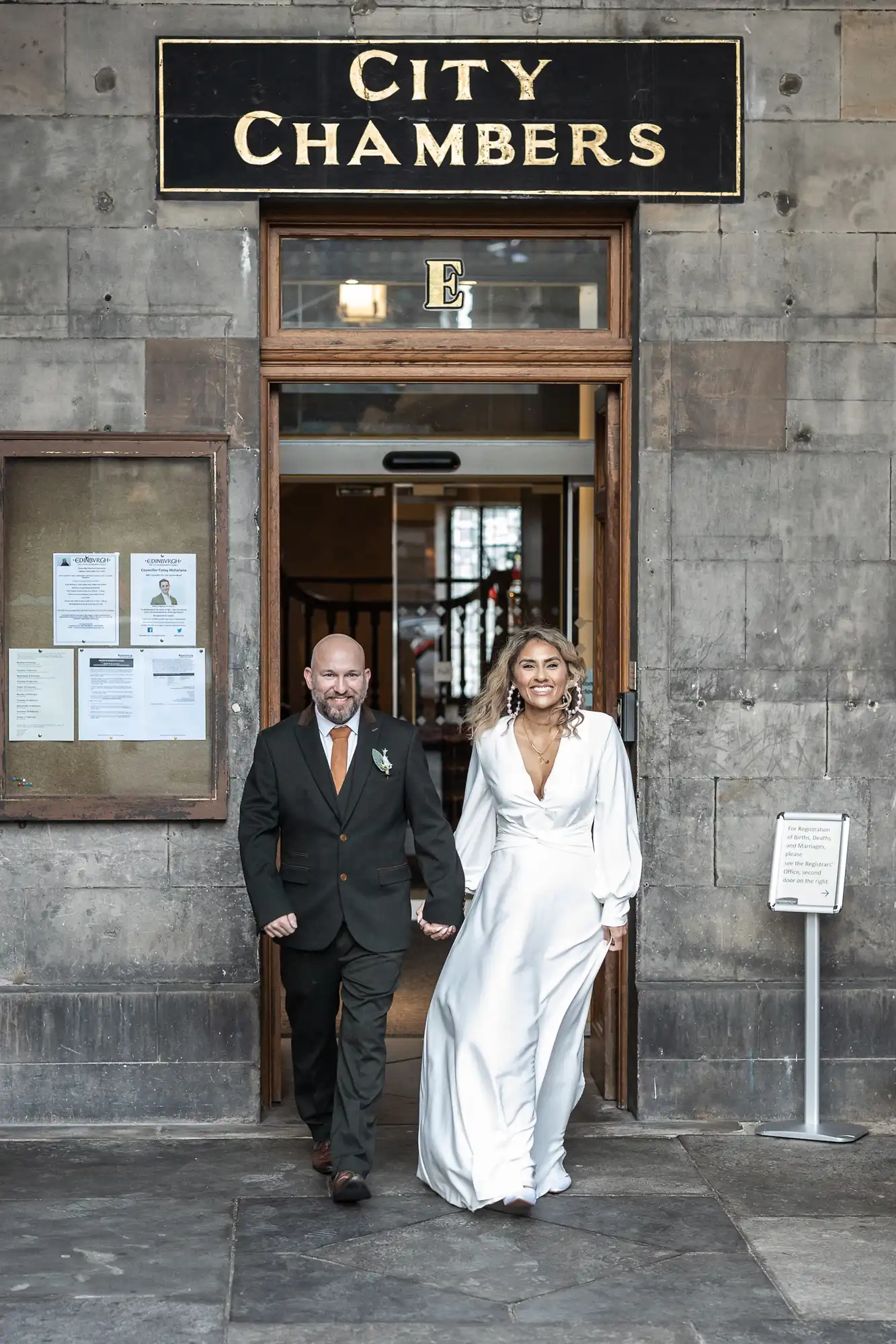 A couple, dressed formally, walks out of a City Chambers building through an open doorway while holding hands, with the woman wearing a white gown and the man in a dark suit.