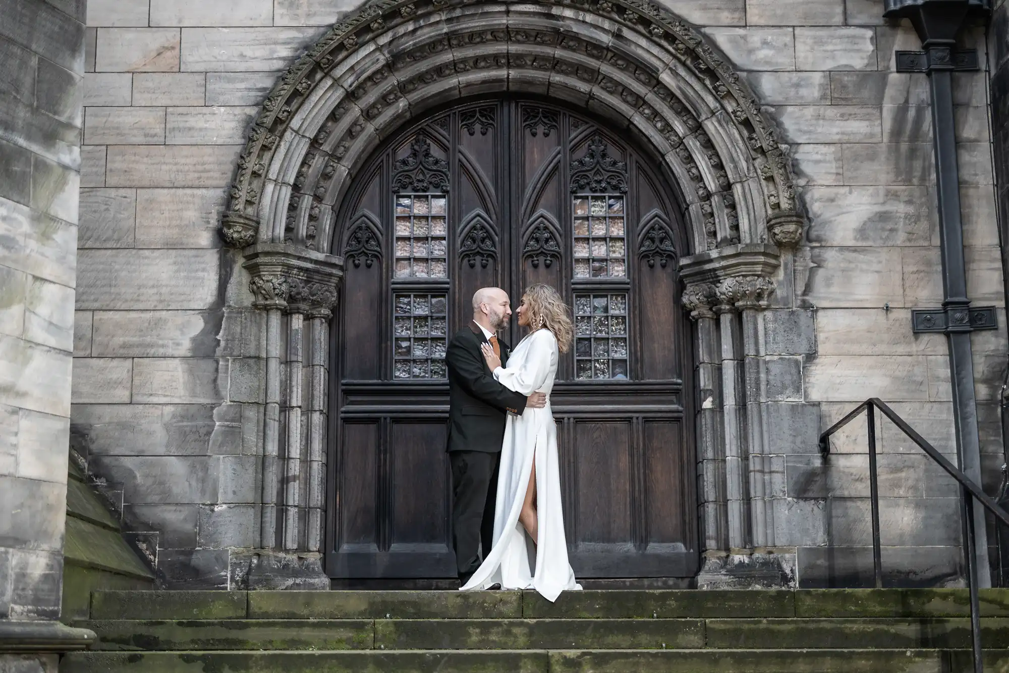 A couple stands closely, looking at one another in front of a large, ornate wooden door with stone arch detailing. The man wears a black suit, and the woman wears a white dress with a slit.