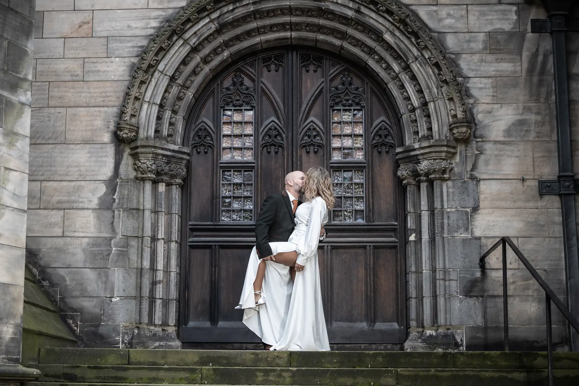 A couple in formal attire kisses in front of a Gothic-style arched wooden door with intricate stone carvings. The man wears a dark suit, and the woman wears a long white dress.