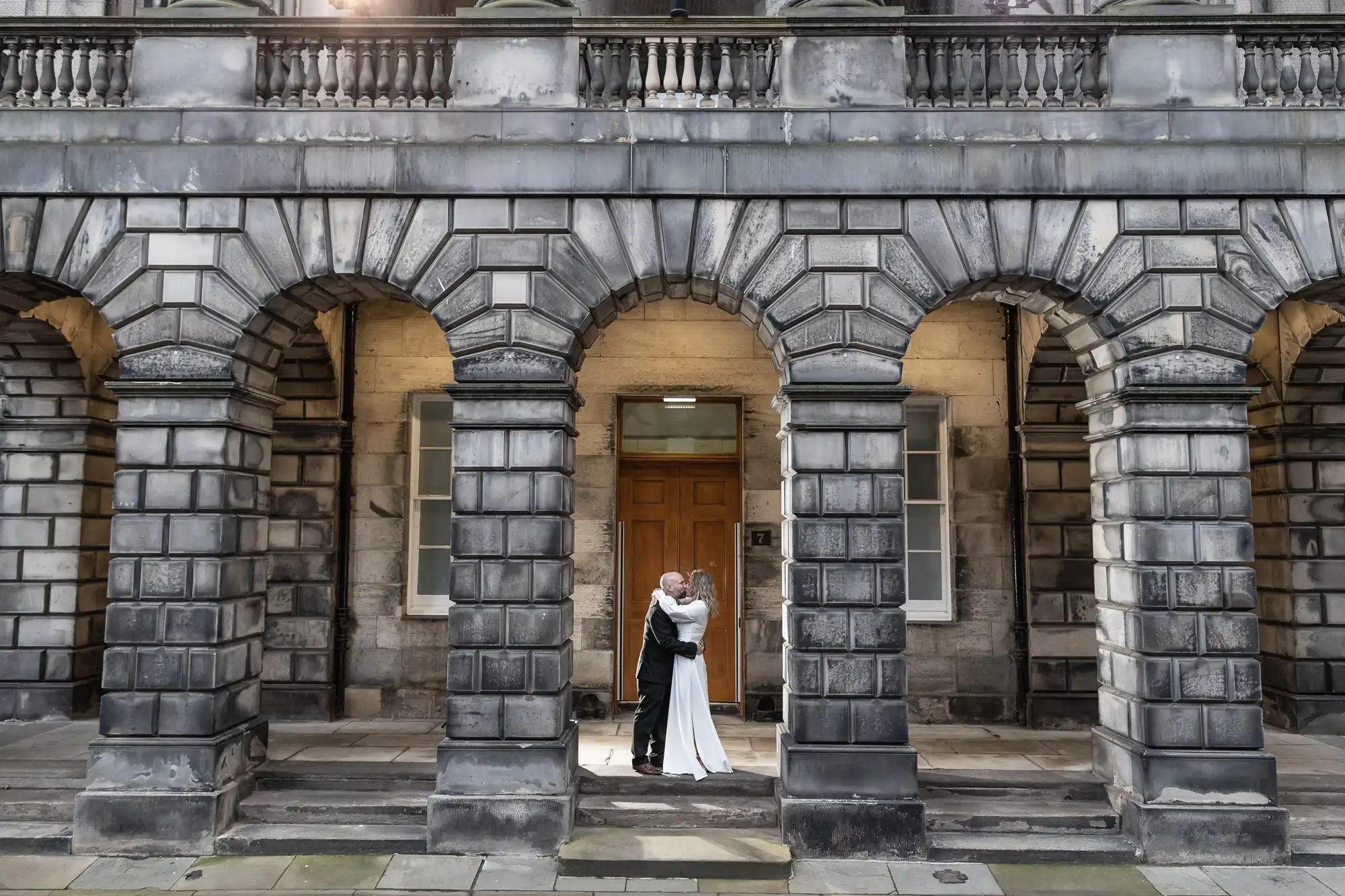 A couple dressed in wedding attire embraces under the archways of a historic stone building.