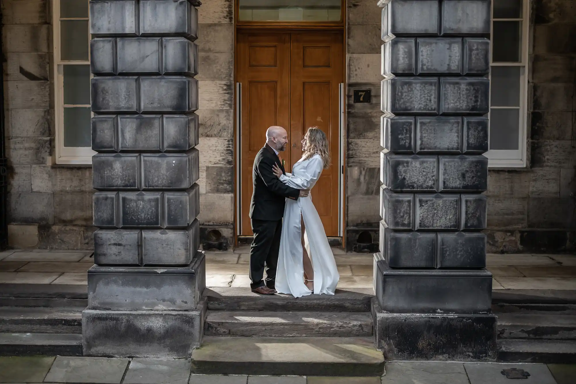 A couple stands facing each other, embrace, and smile between two stone pillars in front of a wooden door. The man wears a suit, and the woman is dressed in a long white outfit.