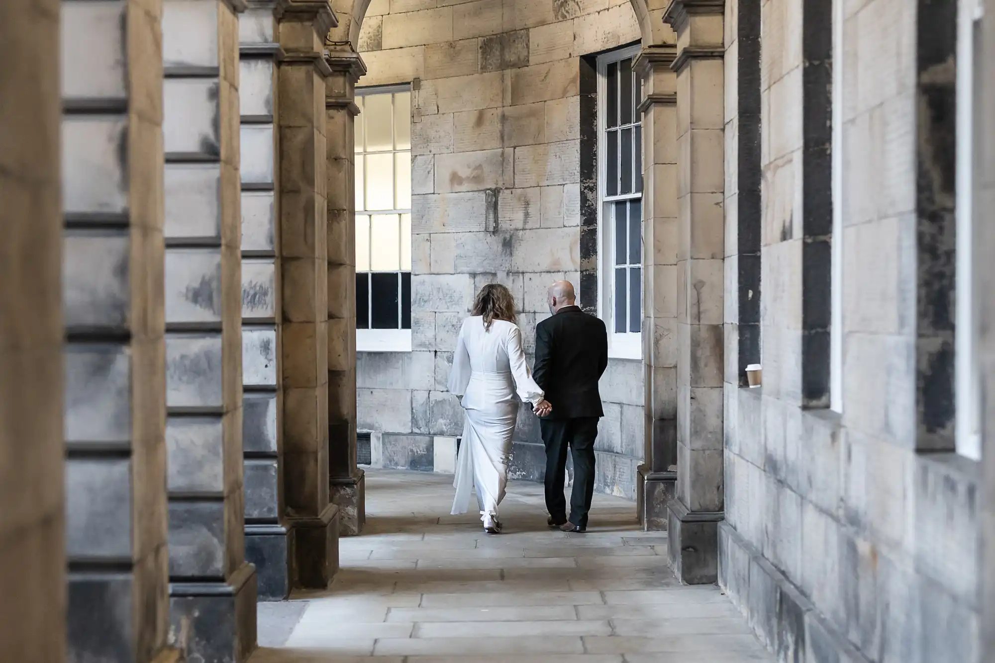 A couple, dressed in formal attire, walks hand-in-hand down a stone hallway, with high arches and large windows.
