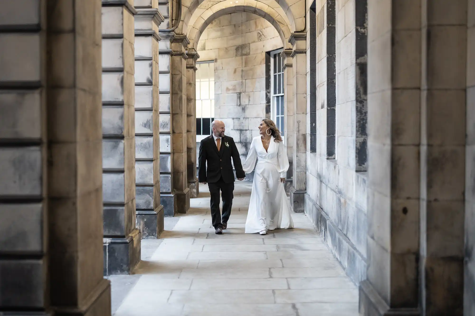 A couple, dressed in wedding attire, walks through a stone archway holding hands. The bride wears a white dress, and the groom wears a dark suit.