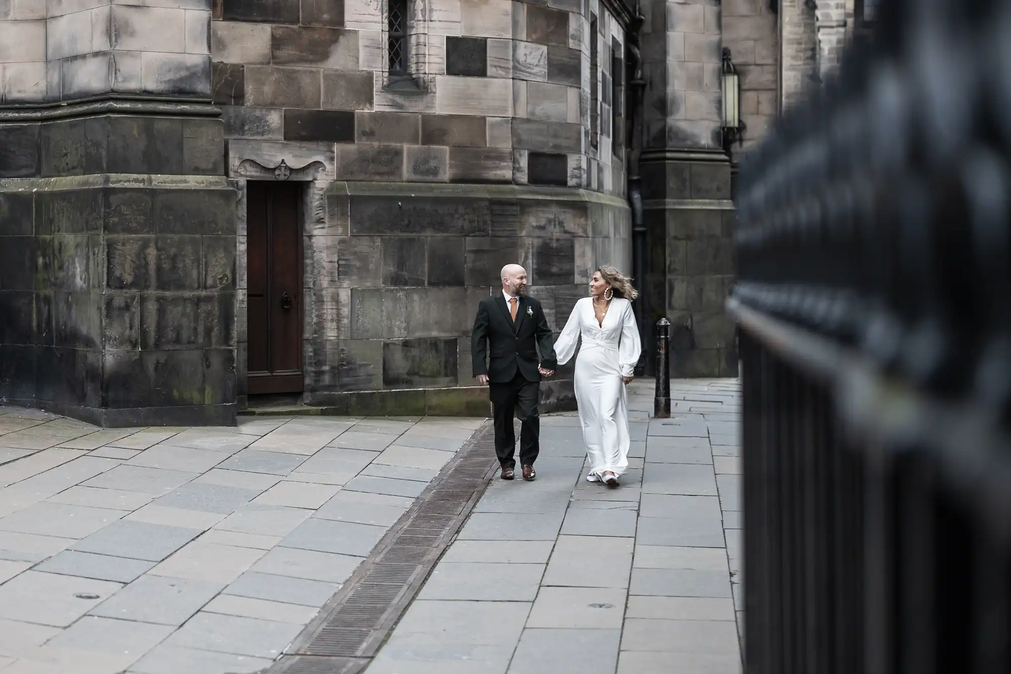 A man in a dark suit and a woman in a white outfit walk hand in hand on a cobblestone street near a stone building.