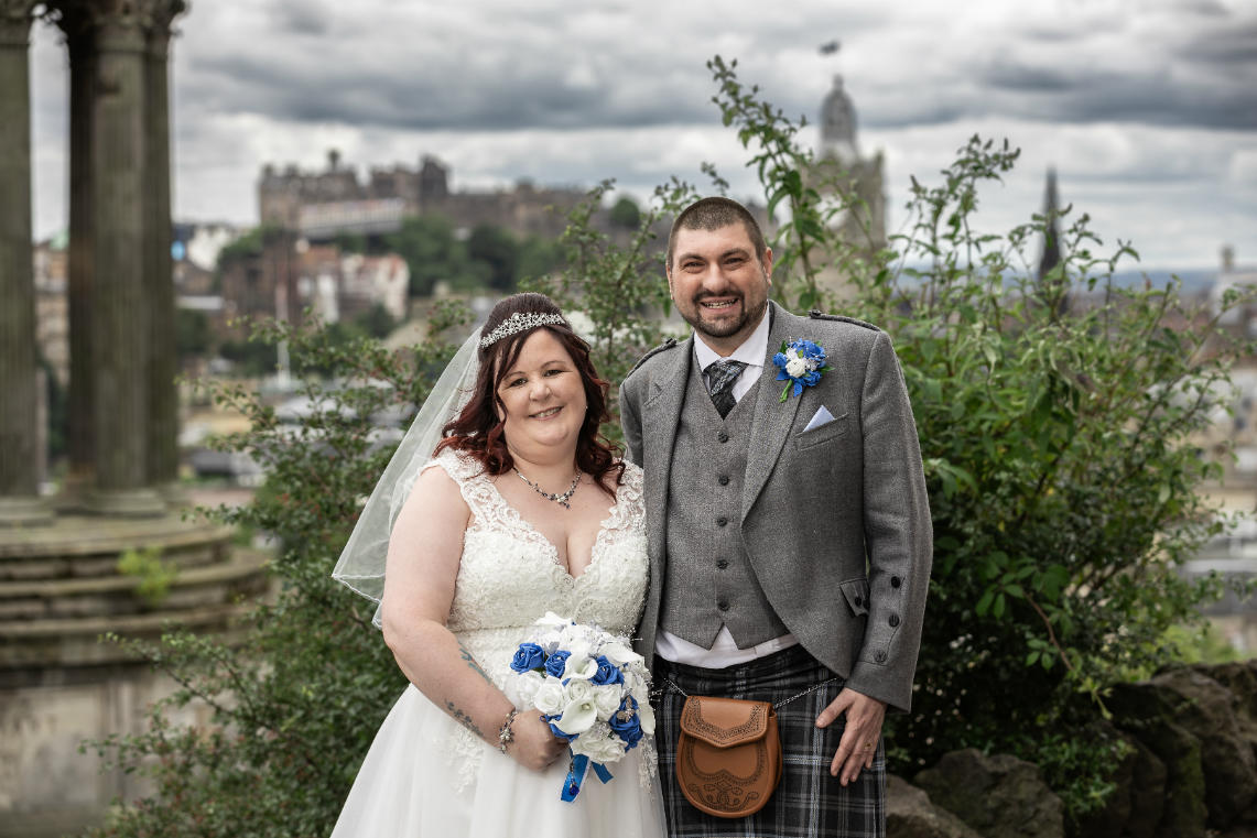 A bride and groom pose outdoors in wedding attire, with greenery and a cityscape featuring historic buildings in the background.