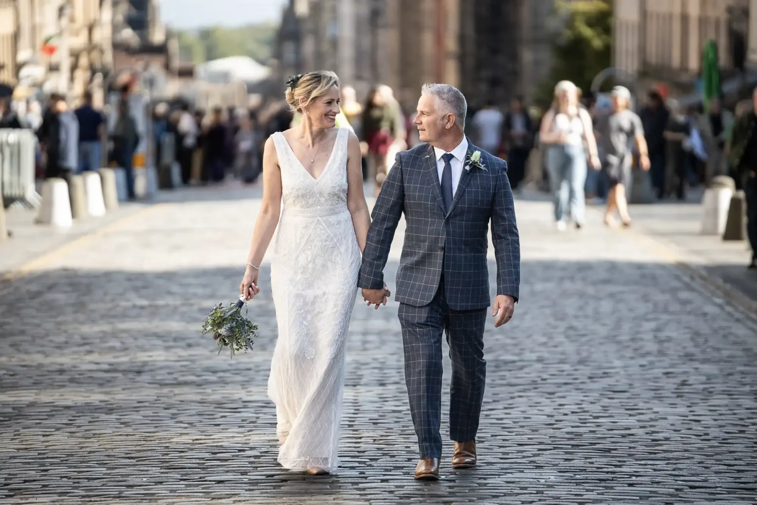 A bride and groom hold hands and walk down a cobblestone street, smiling at each other, with people in the background.