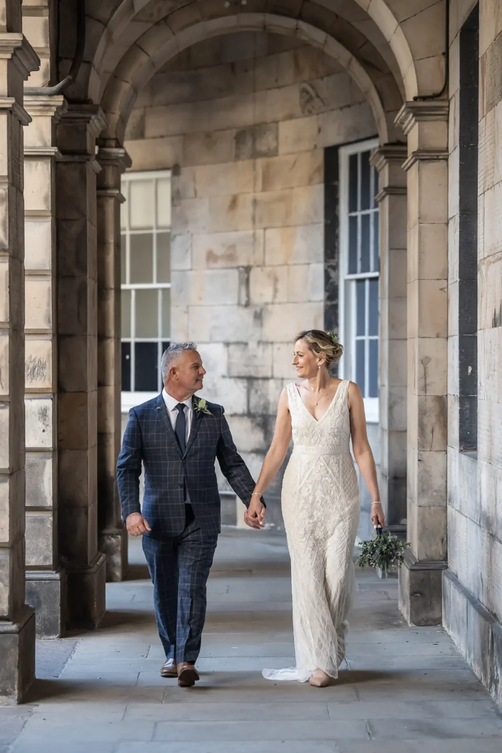 A couple dressed in wedding attire walks hand in hand through a stone archway corridor.