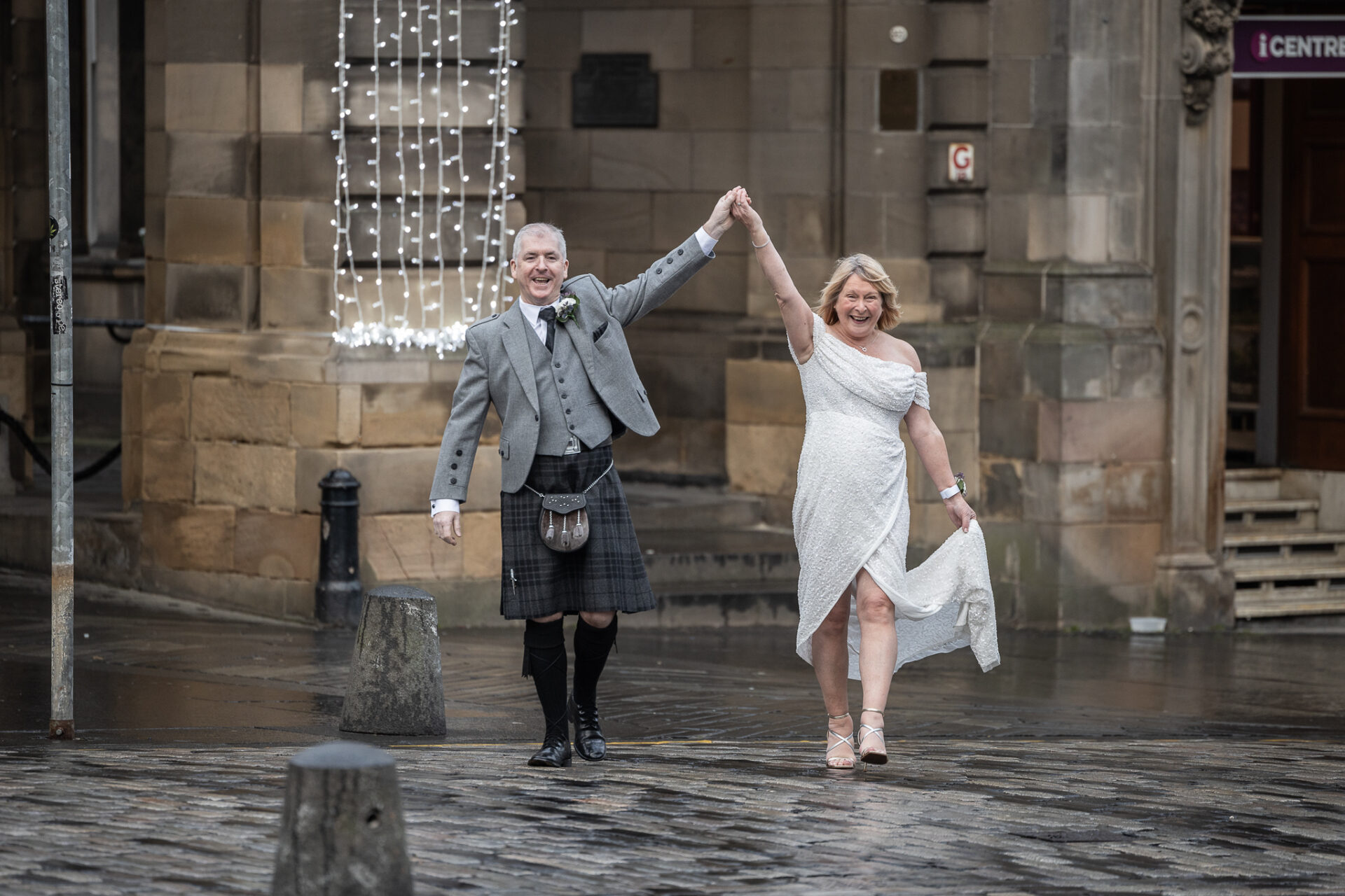 A couple in formal attire joyfully holding hands while walking on a cobblestone street.