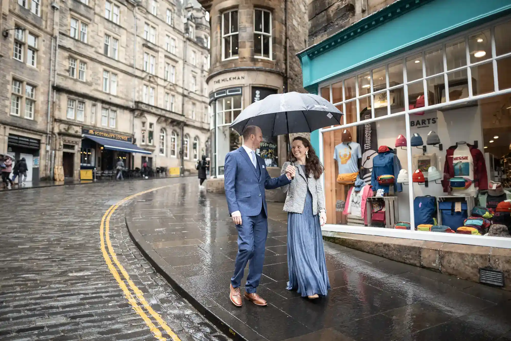A smartly dressed couple walks on a wet cobblestone street, sharing an umbrella near a shop window with colorful bags, in a historic city setting.