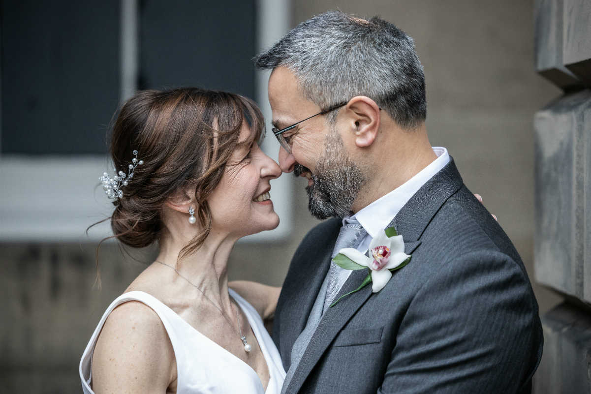 A bride and groom stand close together, smiling and touching noses. The groom wears a dark suit with a boutonniere, and the bride wears a white dress with a hair accessory.