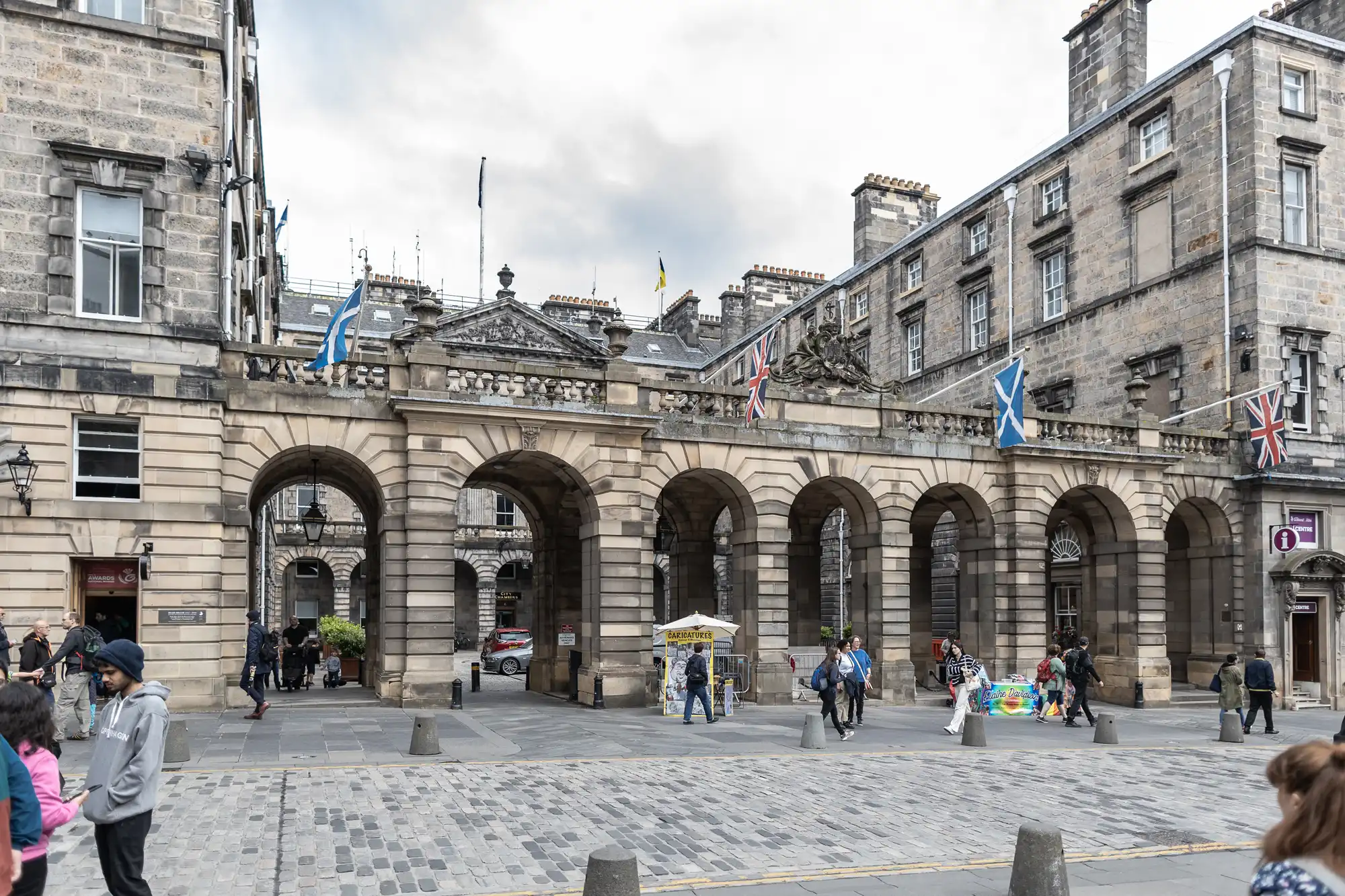 Historic stone building with arches, adorned with Scottish flags and Union Jacks, situated on a cobblestone street with pedestrians walking and street vendors visible.