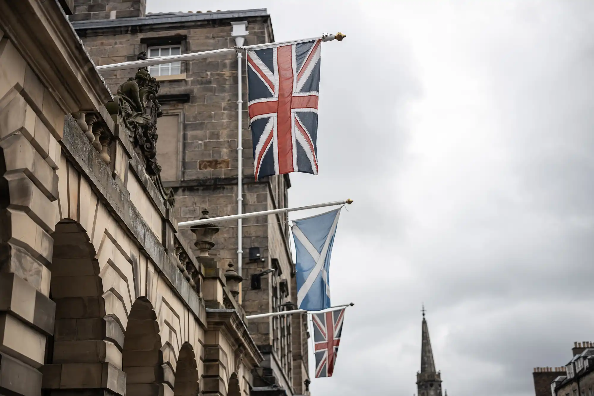 Flags of the United Kingdom and Scotland are displayed on flagpoles attached to a stone building, with a cloudy sky and a tower visible in the background.