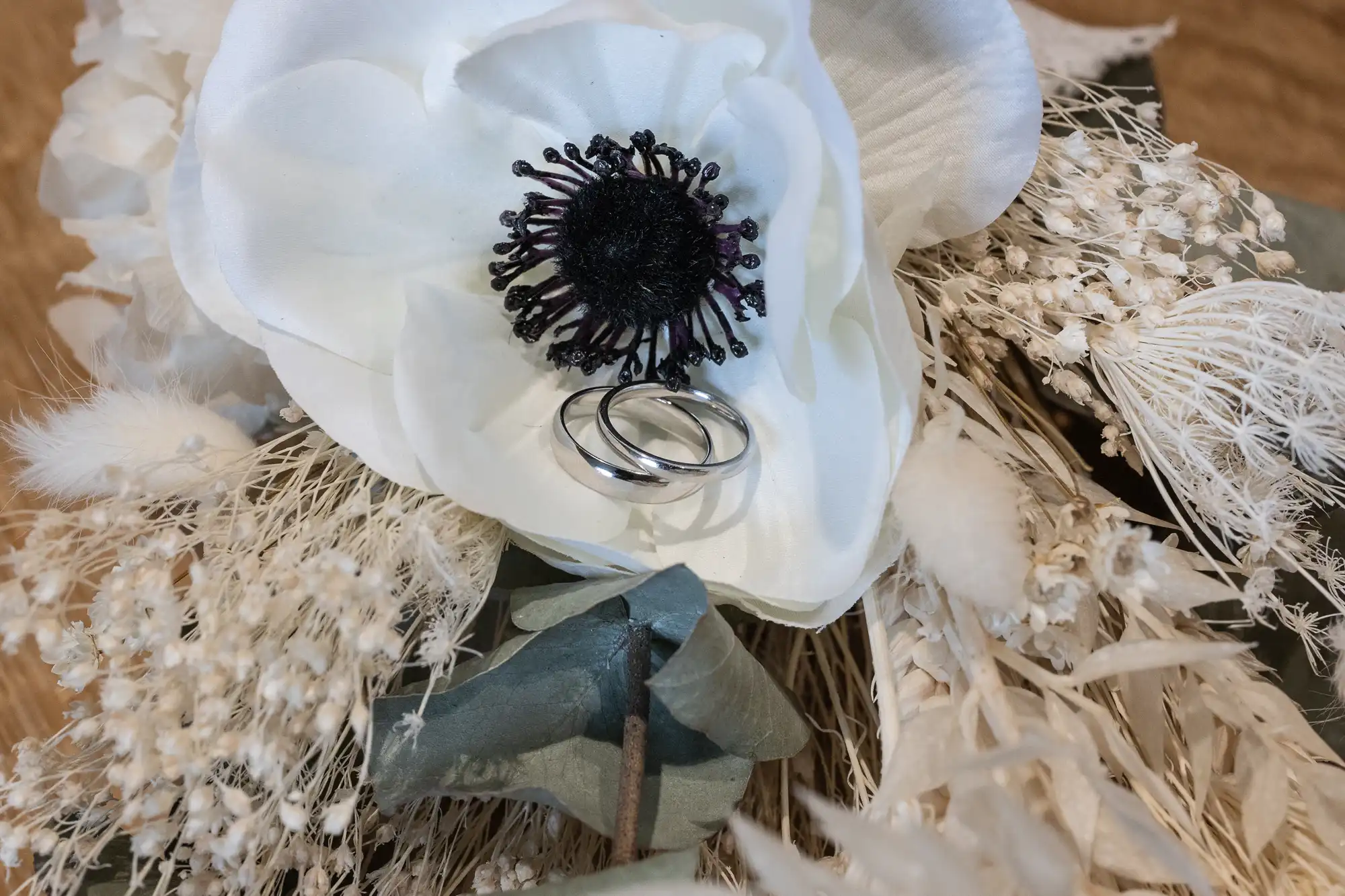 Two silver wedding rings rest on a white flower with black center, surrounded by an arrangement of dried flowers and grasses.