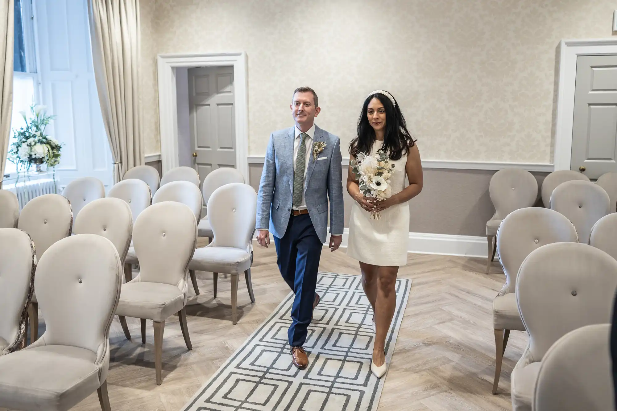 A couple walks down the aisle in a small, elegantly decorated room with beige chairs and light walls. The woman holds a bouquet and the man wears a gray jacket with a blue tie.