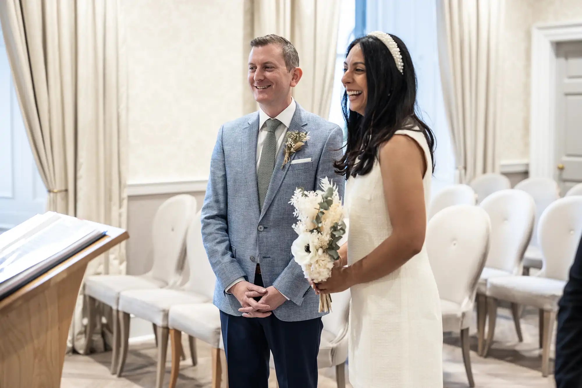 A couple stands together smiling in a small, elegantly decorated room, the woman holding a bouquet of flowers, likely during a civil wedding ceremony.