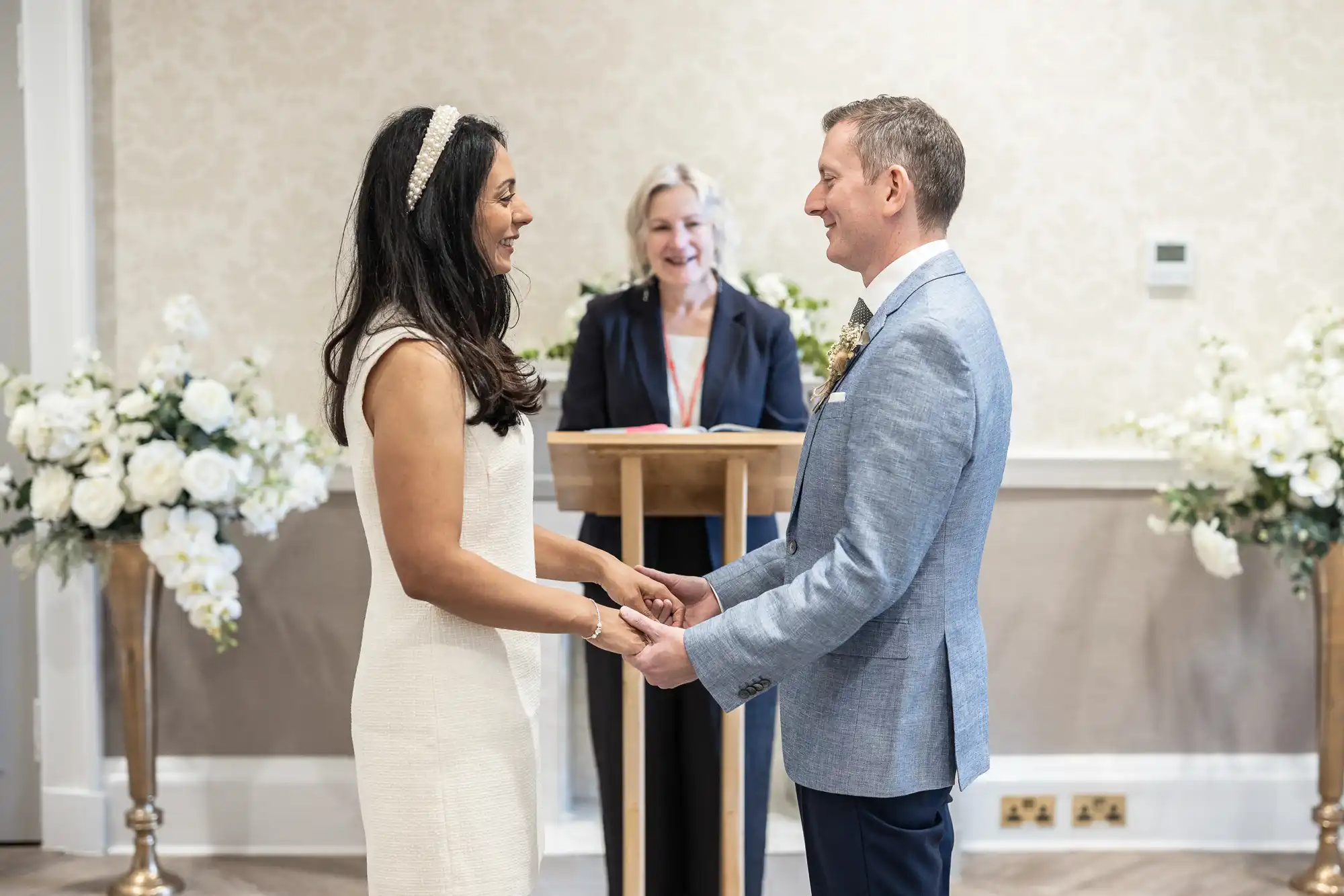 A couple is exchanging vows at their wedding ceremony, standing in front of an officiant. Both are smiling and holding hands. White floral arrangements are displayed on either side of them.