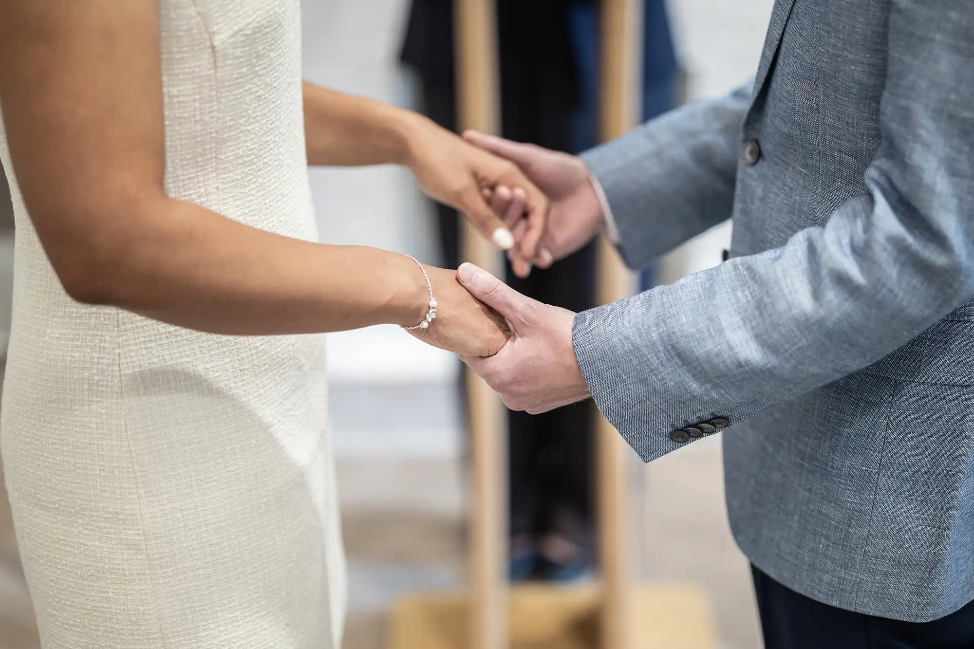 Two people facing each other and holding hands during a wedding ceremony. The person on the left wears a white dress, and the person on the right wears a light gray suit.