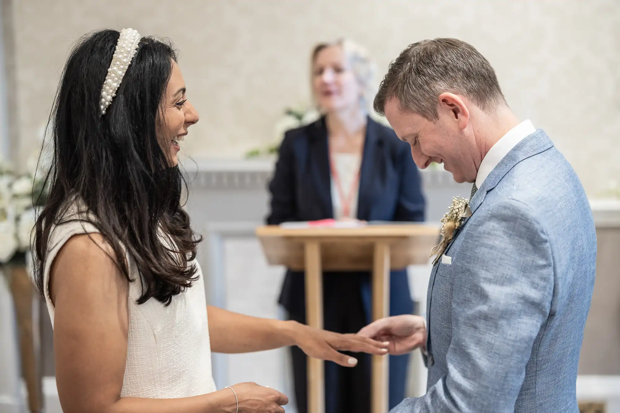 A couple is exchanging rings during a wedding ceremony. An officiant is standing in the background, overseeing the event.