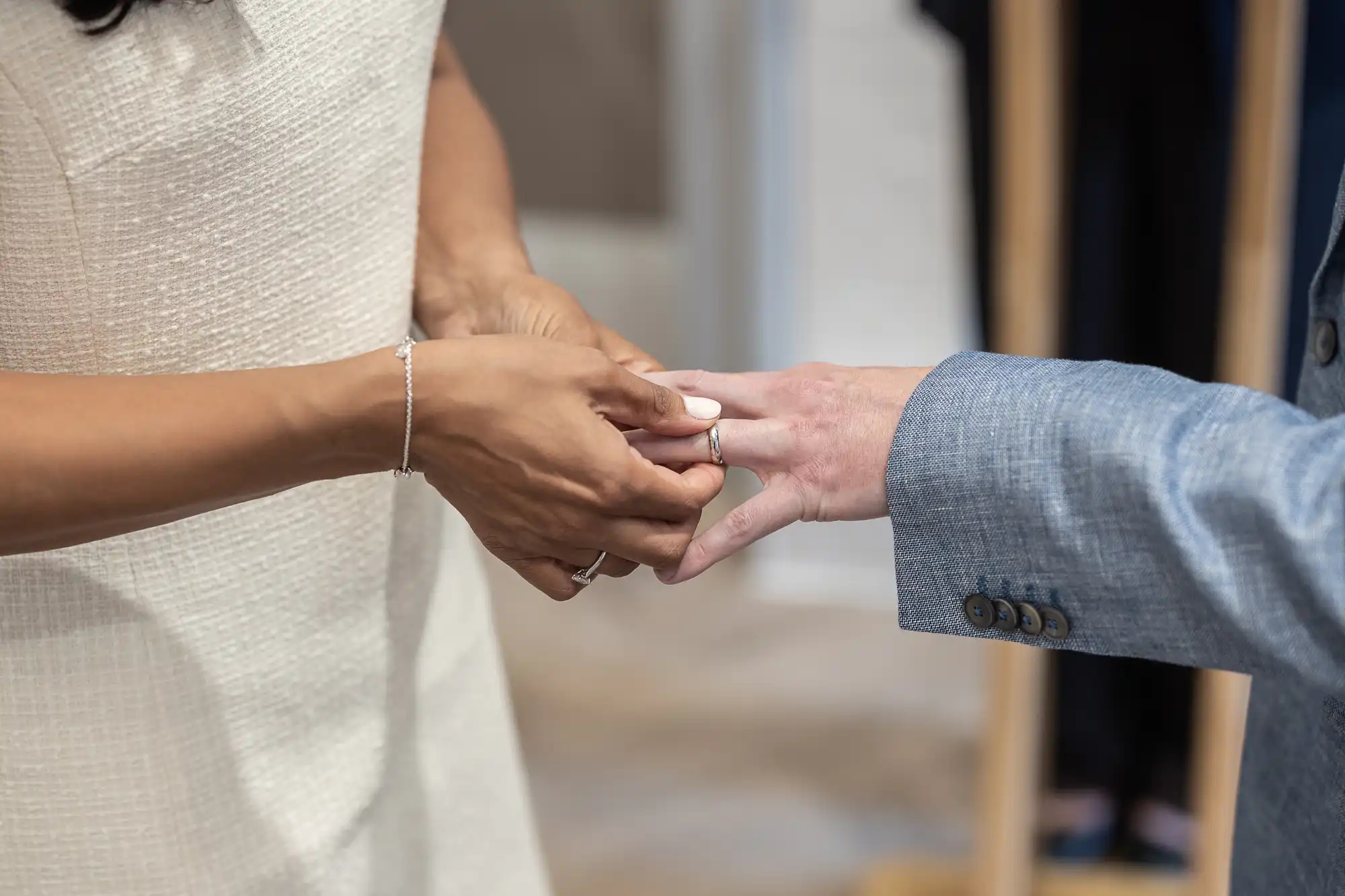 A person in a white dress places a ring on the finger of another person wearing a light blue suit jacket.