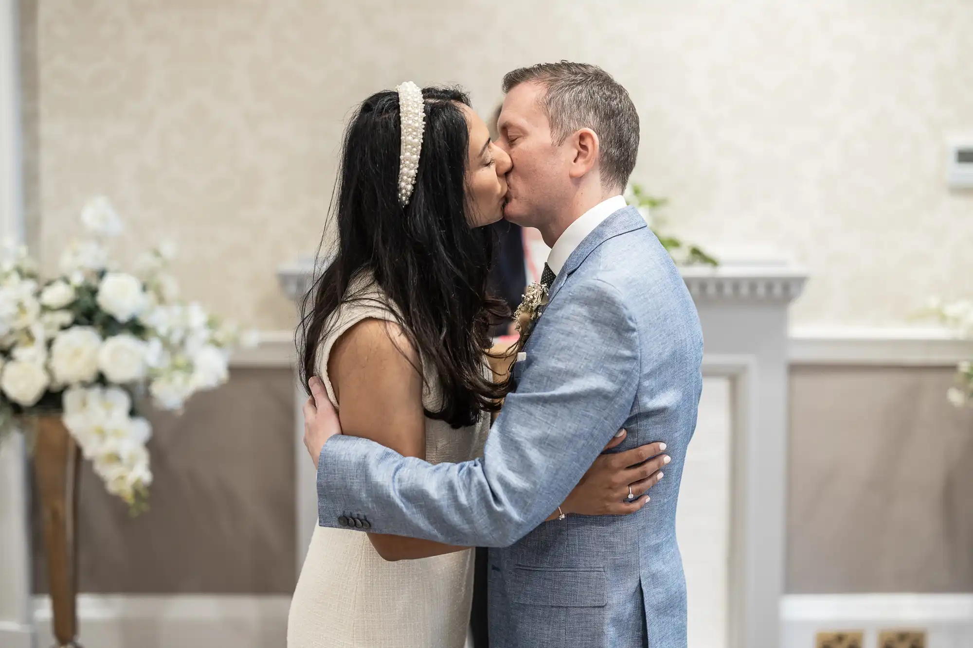 A couple, a woman in a white dress and a man in a light blue suit, share a kiss in a room decorated with white flowers.
