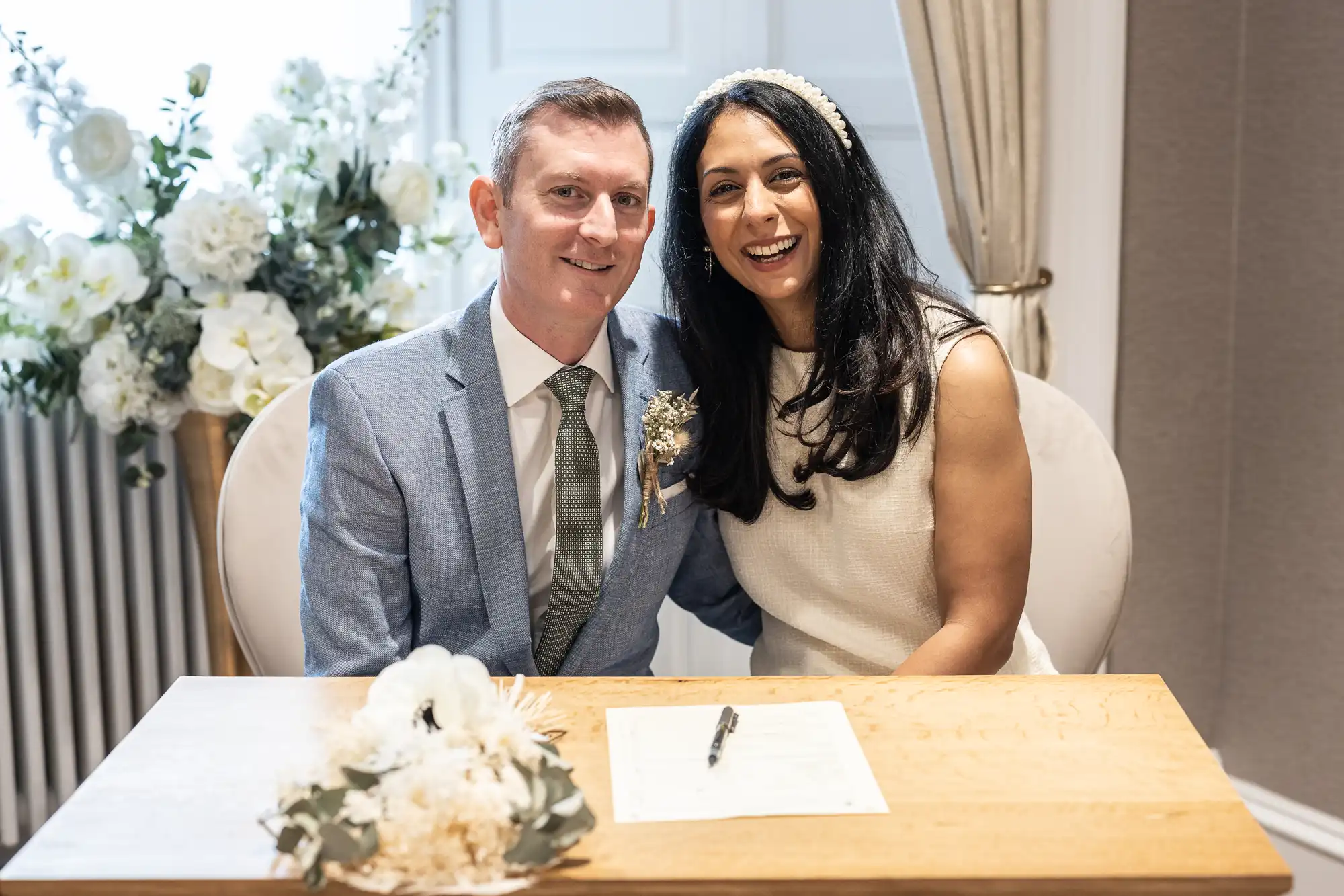 A man in a light gray suit and a woman in a white dress sit together at a table with a paper and pen, smiling. White floral decorations are in the background.