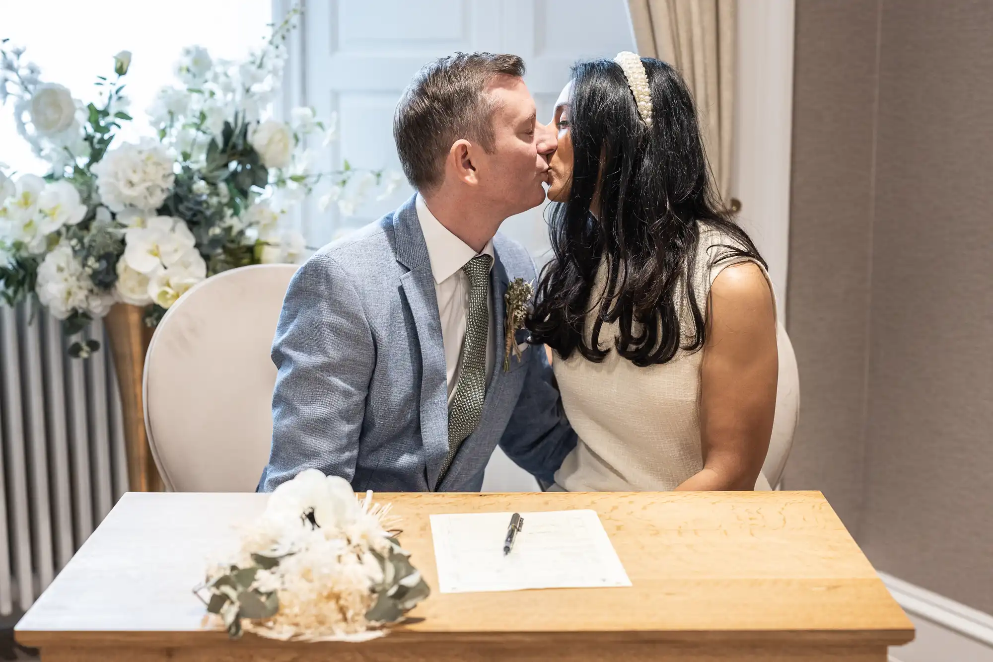 A couple kisses while seated at a wooden table with a document and pen. A bouquet of flowers is on the table. A floral arrangement is visible in the background.