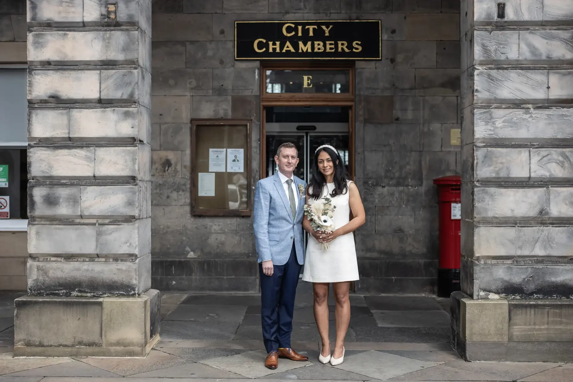 wedding photographer Edinburgh City Chambers - a couple, dressed formally, stands in front of the City Chambers building entrance, with the woman holding a bouquet of flowers.